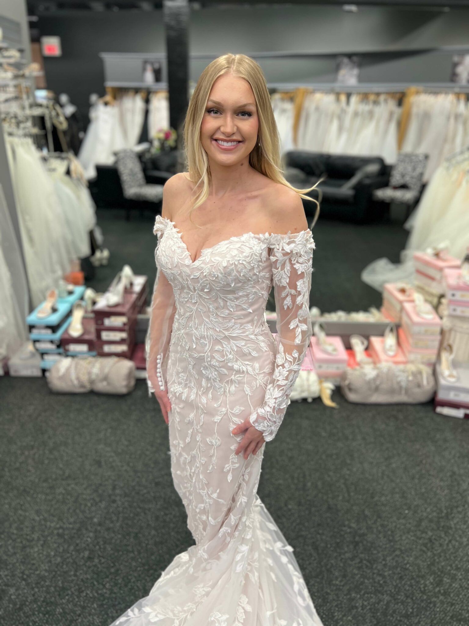 Woman in a white off-shoulder wedding dress smiles in a bridal shop.