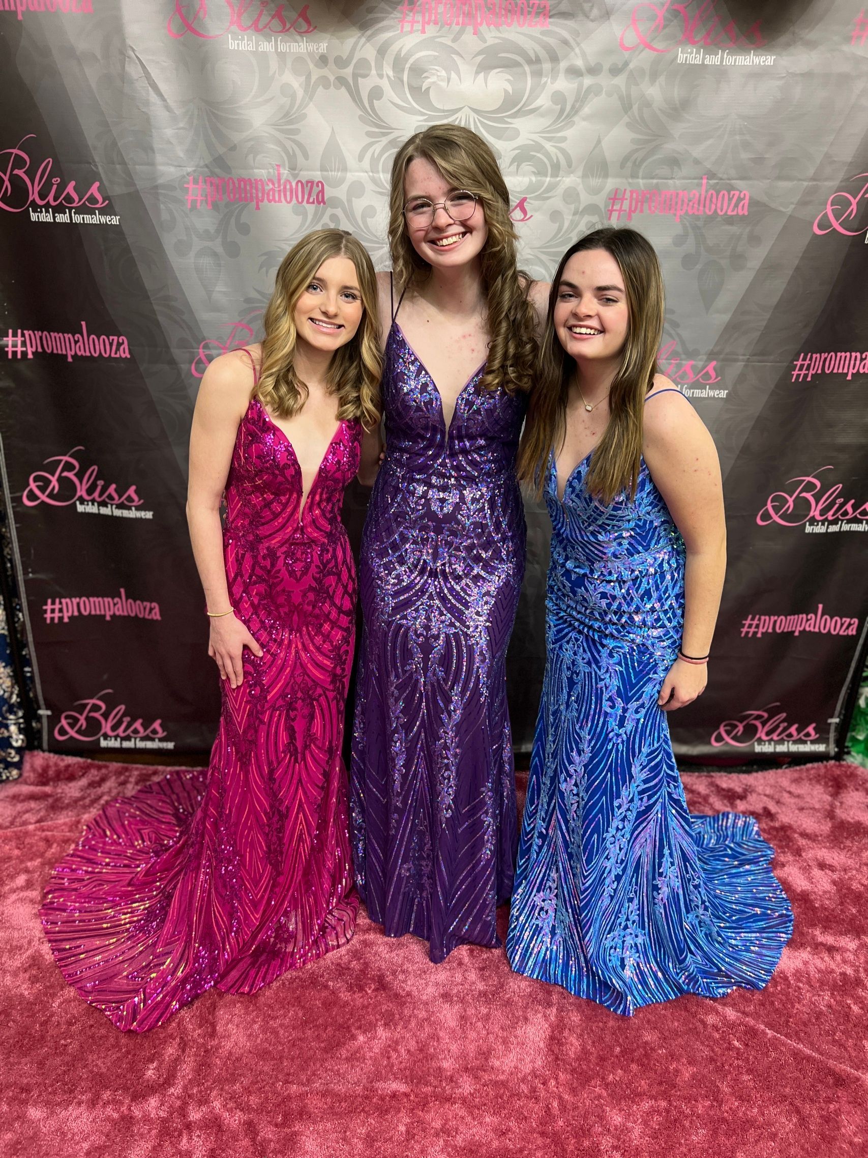 Three young people in formal dresses pose for a photo. A purple carpet and backdrop. Dresses are hot pink, purple, and blue.