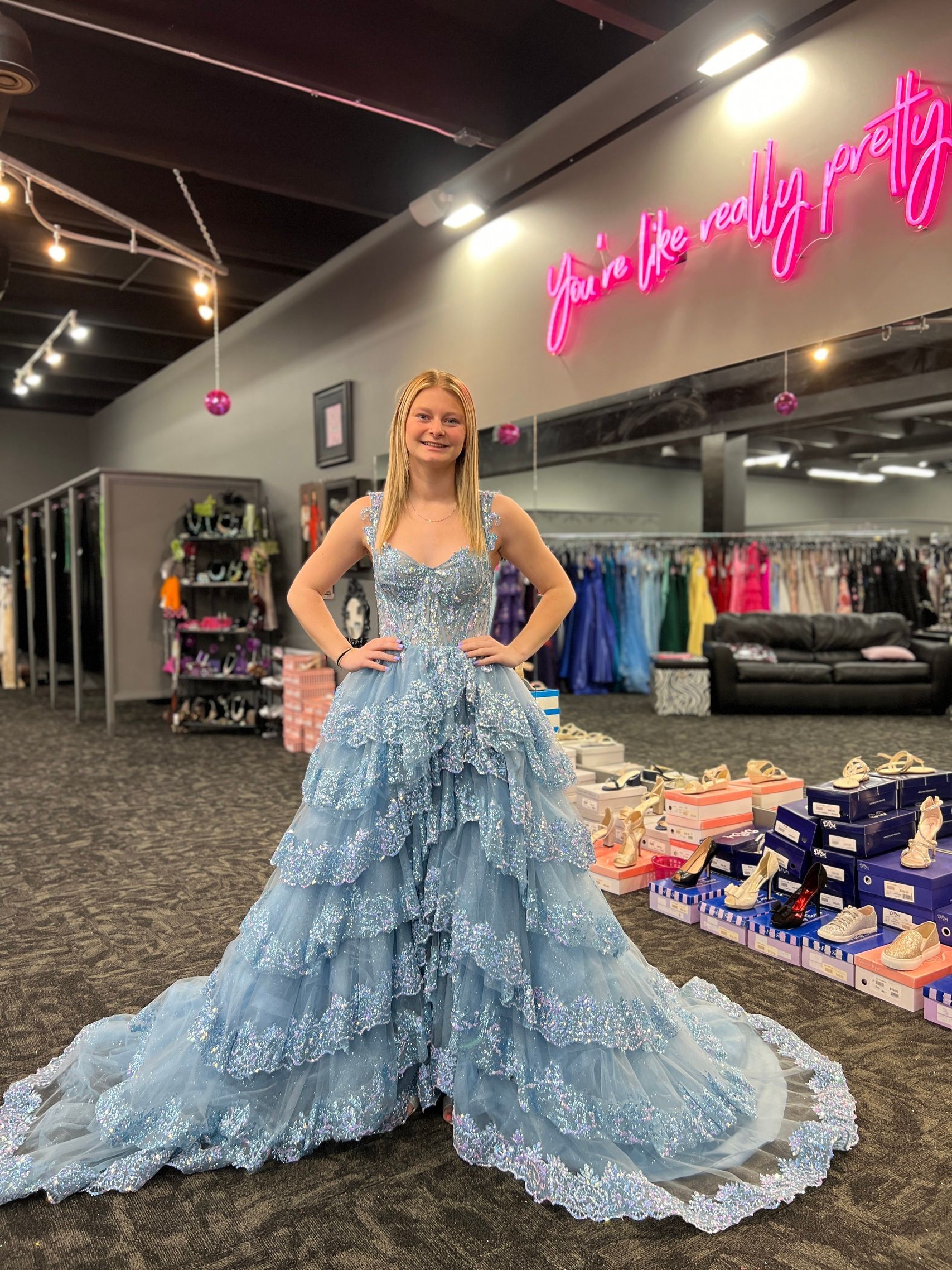 Woman in a blue gown with floral detail poses in a prom dress shop, hands on hips.