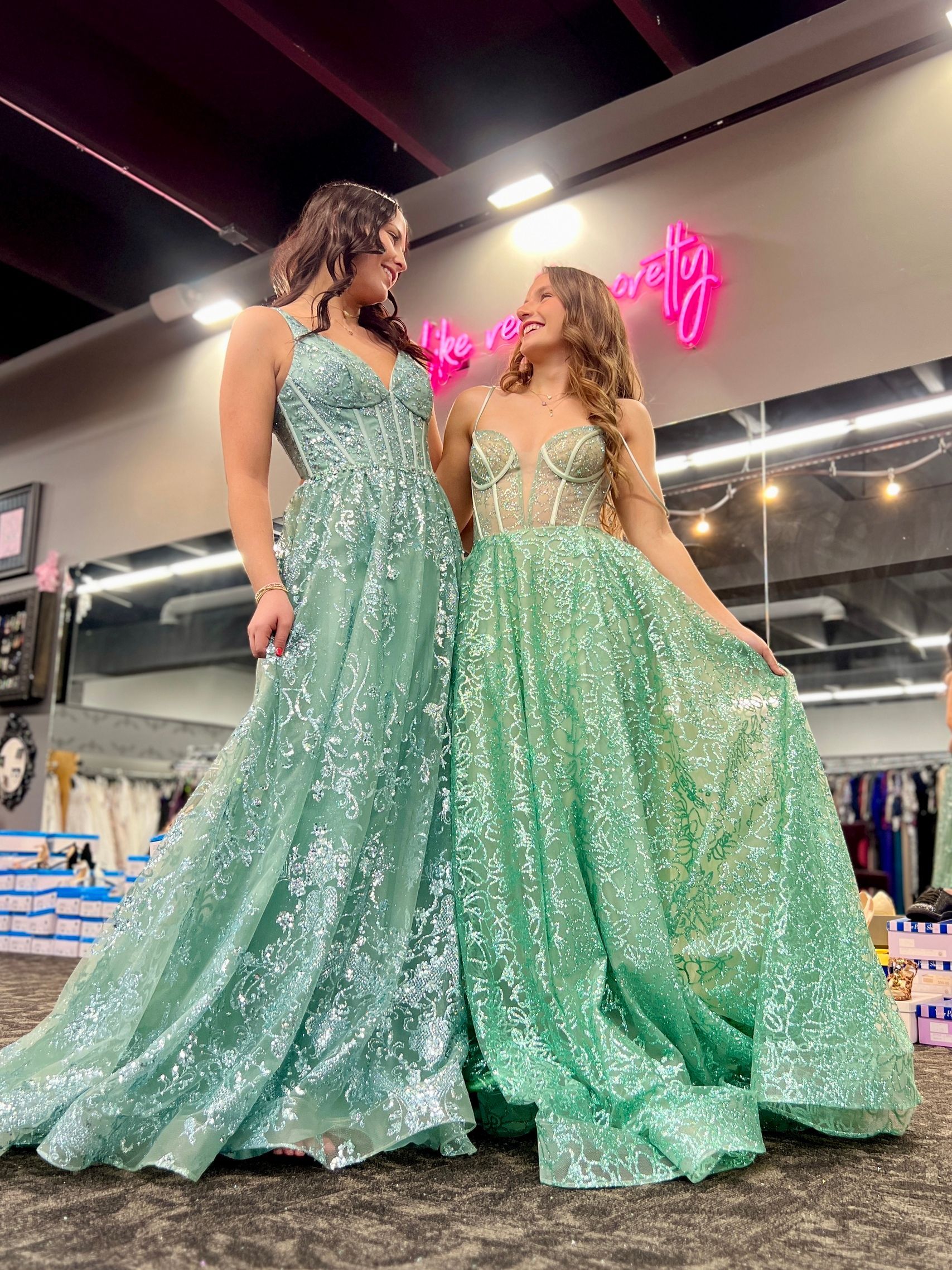 Two women in sparkly, mint green formal gowns, standing and smiling in a dress shop.