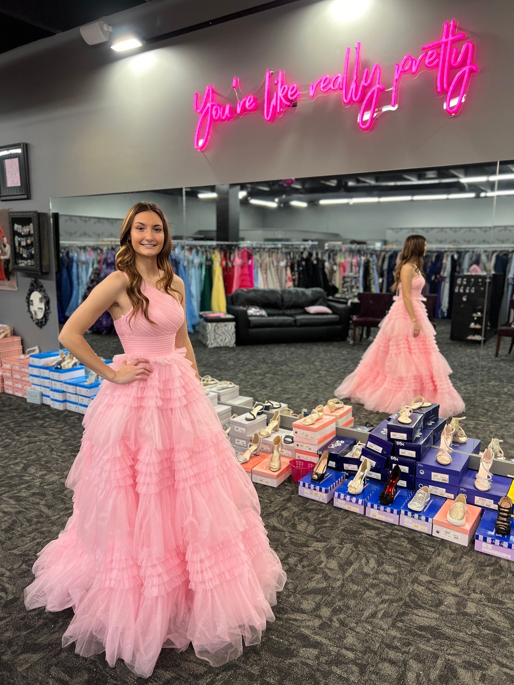 Woman in pink gown poses in a dress shop. A second woman stands in the background wearing the same gown.