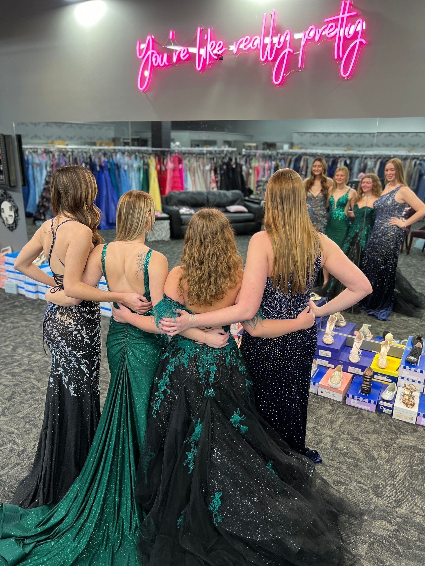 Five women in prom dresses pose arm-in-arm in a dress shop under a neon sign that says