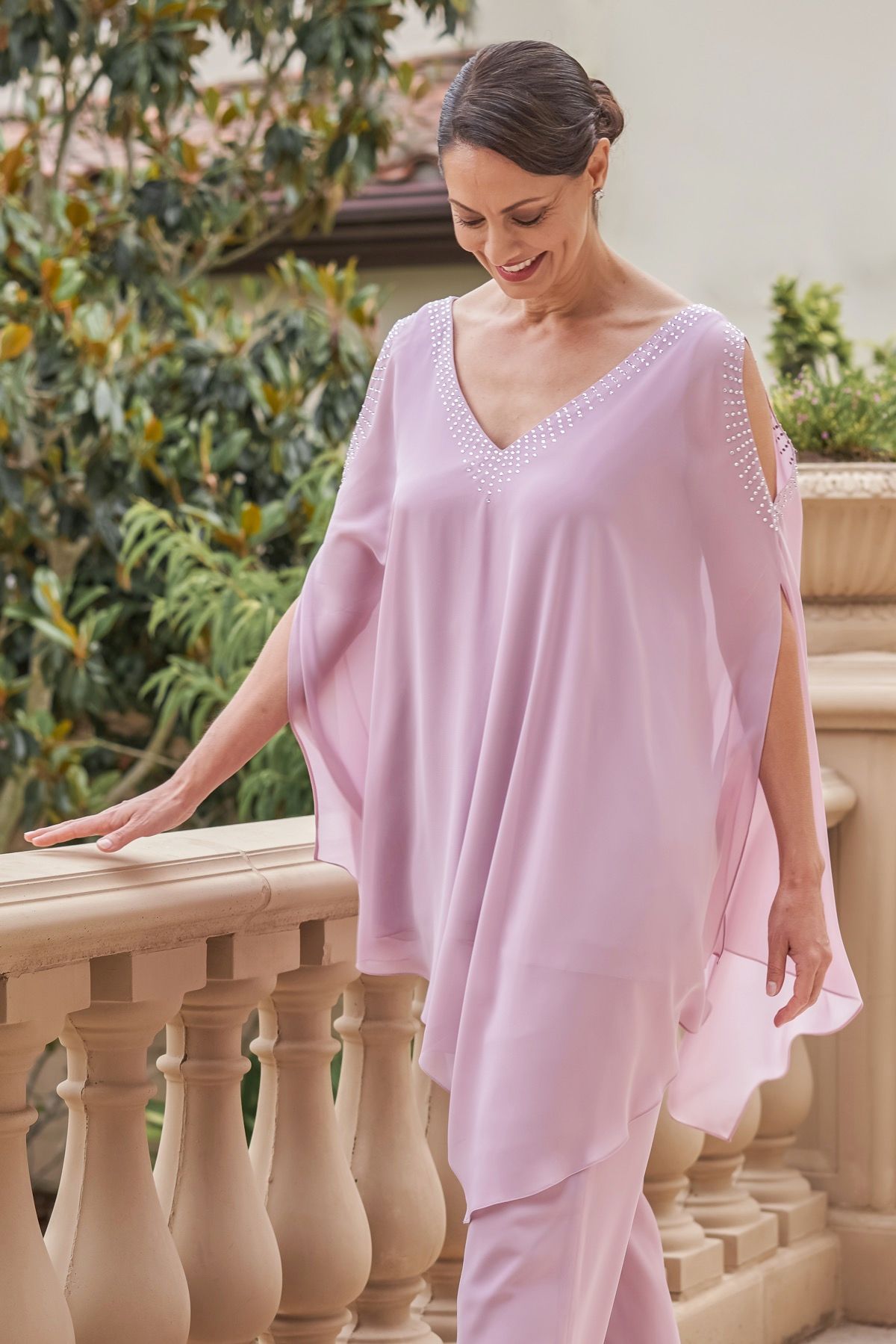 Woman in pink dress with jeweled neckline smiles, leans on stone balcony, outdoor setting.
