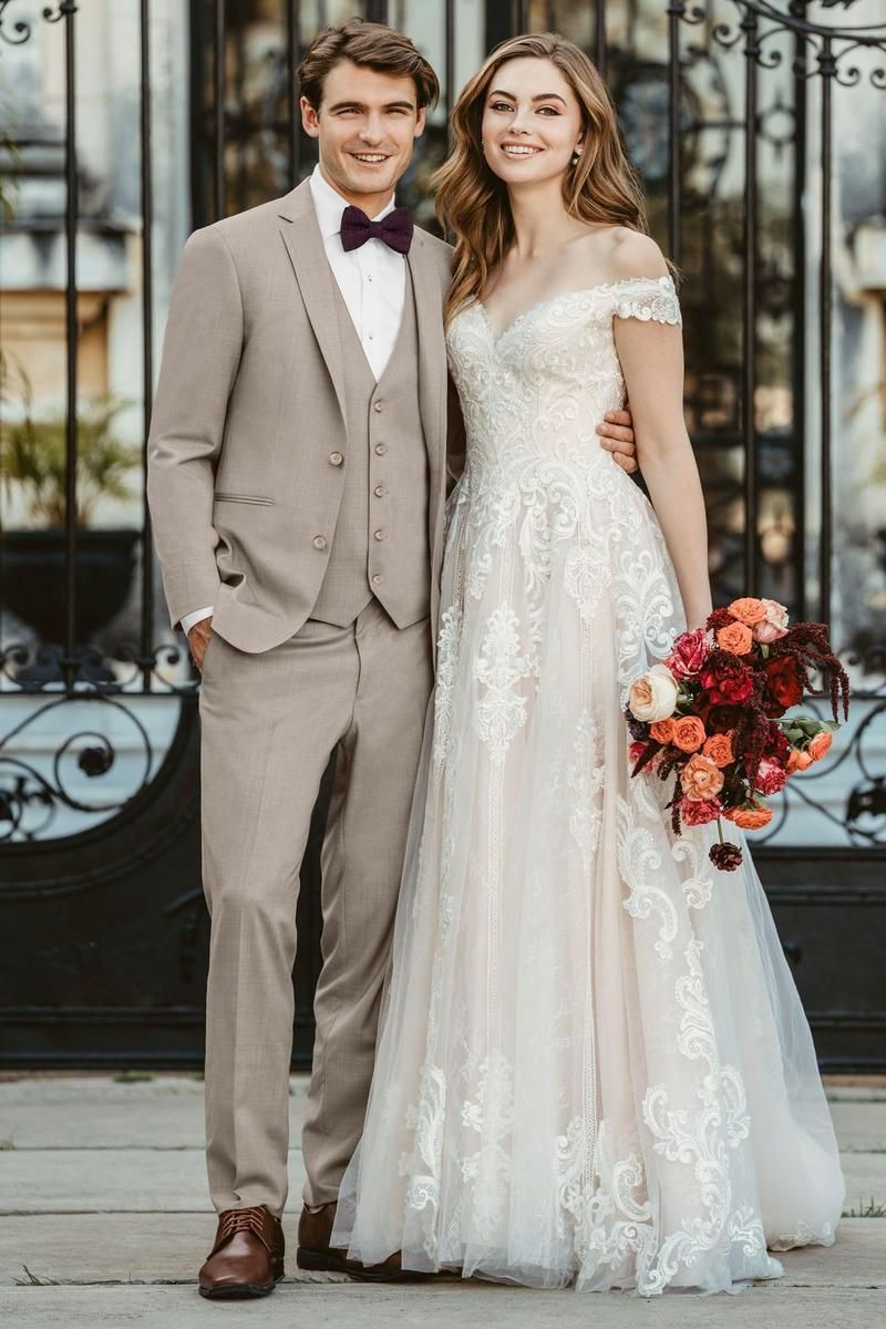 Couple in wedding attire posing outdoors. Man in tan suit, woman in off-the-shoulder dress, holding flowers.