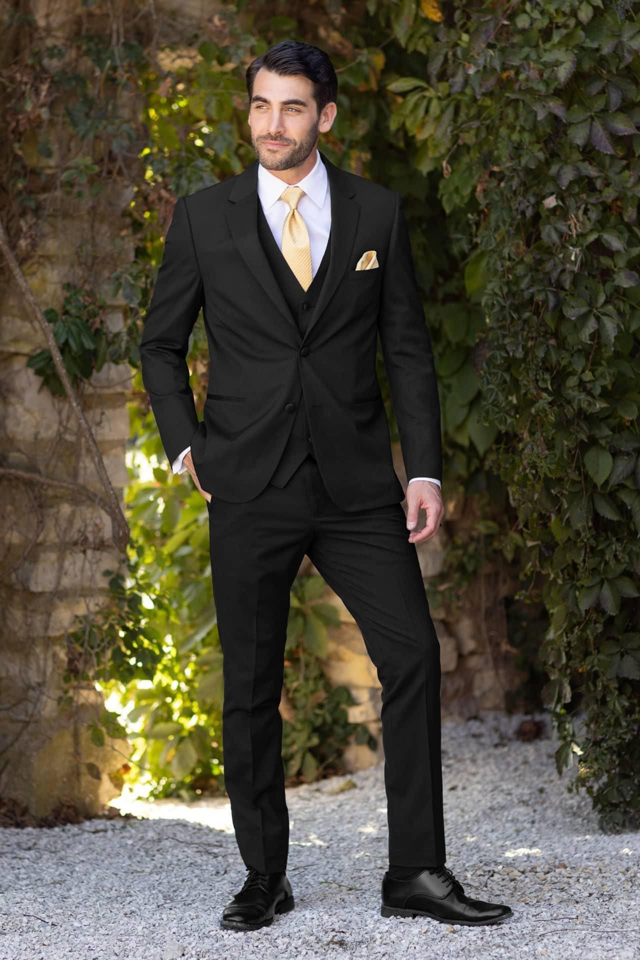 Man in a black suit with a yellow tie and pocket square, standing outdoors by ivy-covered wall.