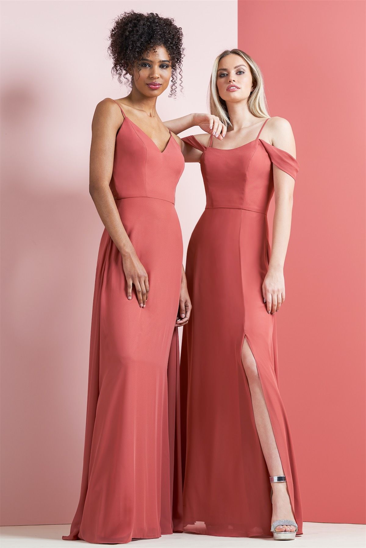 Two women in terracotta-colored bridesmaid dresses, posing next to a coral wall.