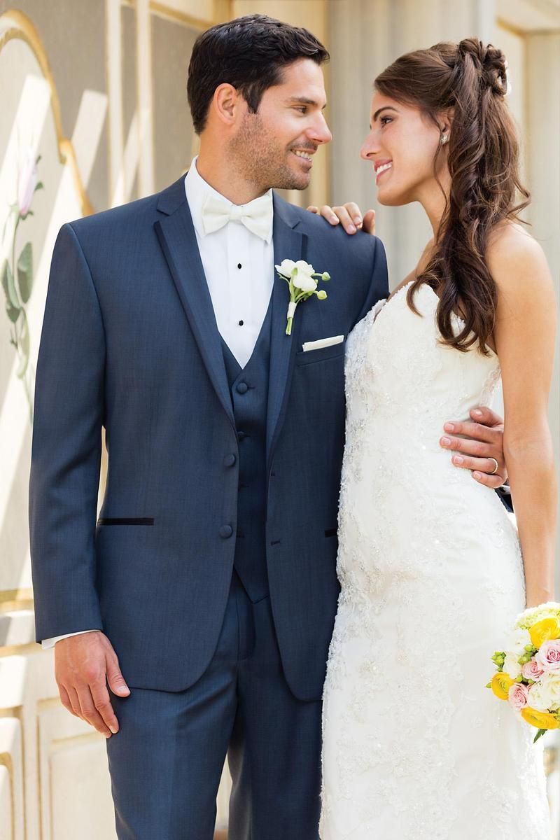 Groom in navy tuxedo with bride in white gown, smiling at each other. Outdoor setting.
