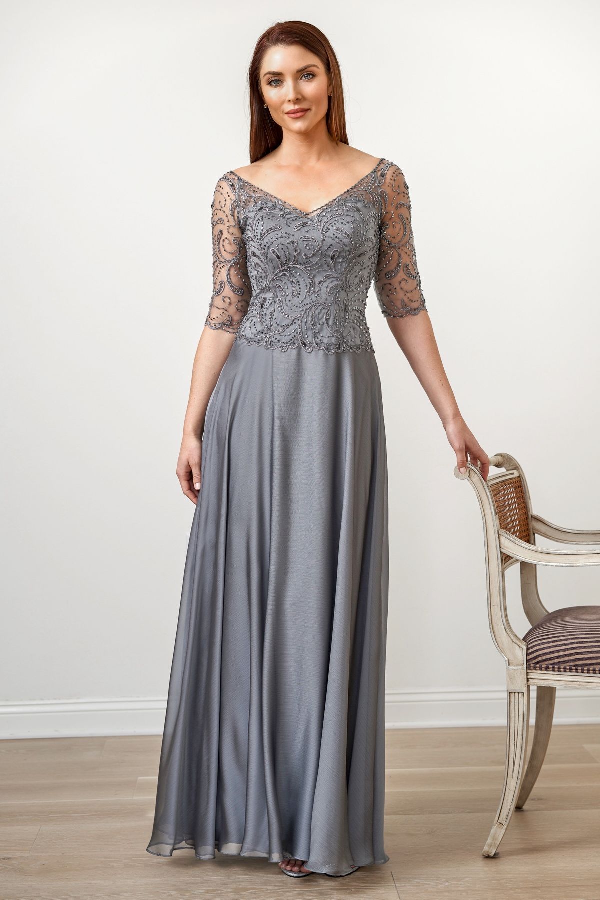 Woman in gray evening gown, beaded bodice, 3/4 sleeves, standing near chair, neutral setting.
