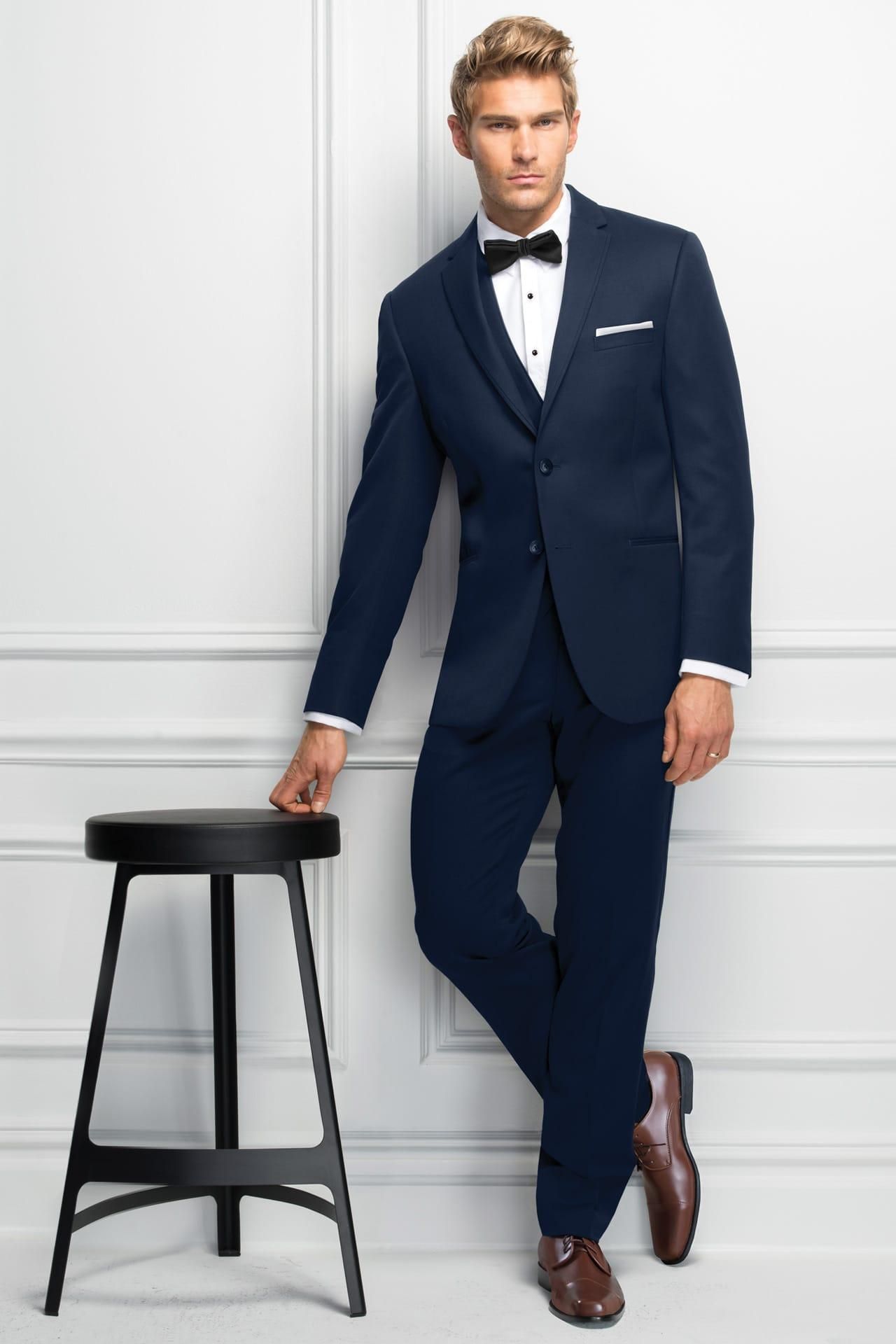 Man in navy suit, white shirt, black bow tie, leaning on stool.