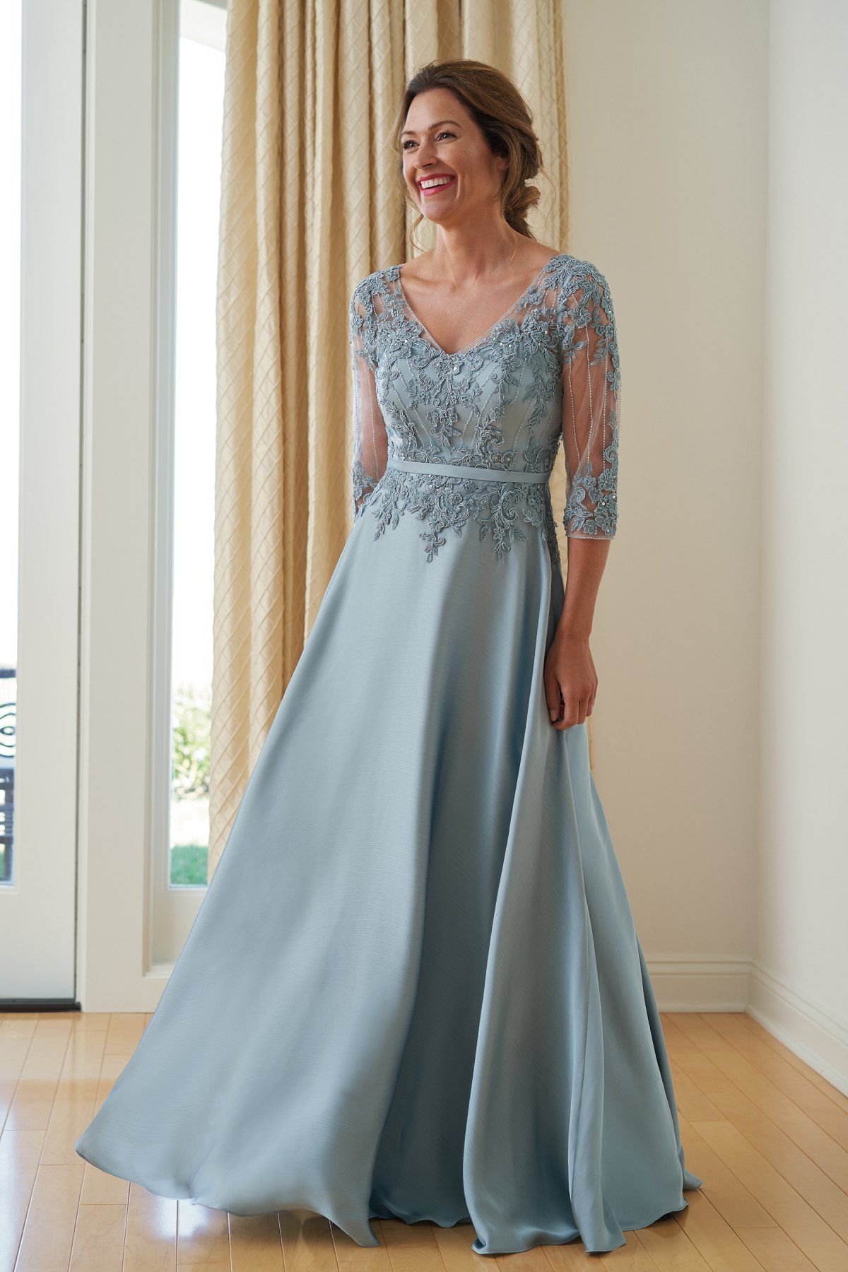 Woman in a light blue formal gown smiles near a window. The dress has lace sleeves and bodice, flowing skirt.