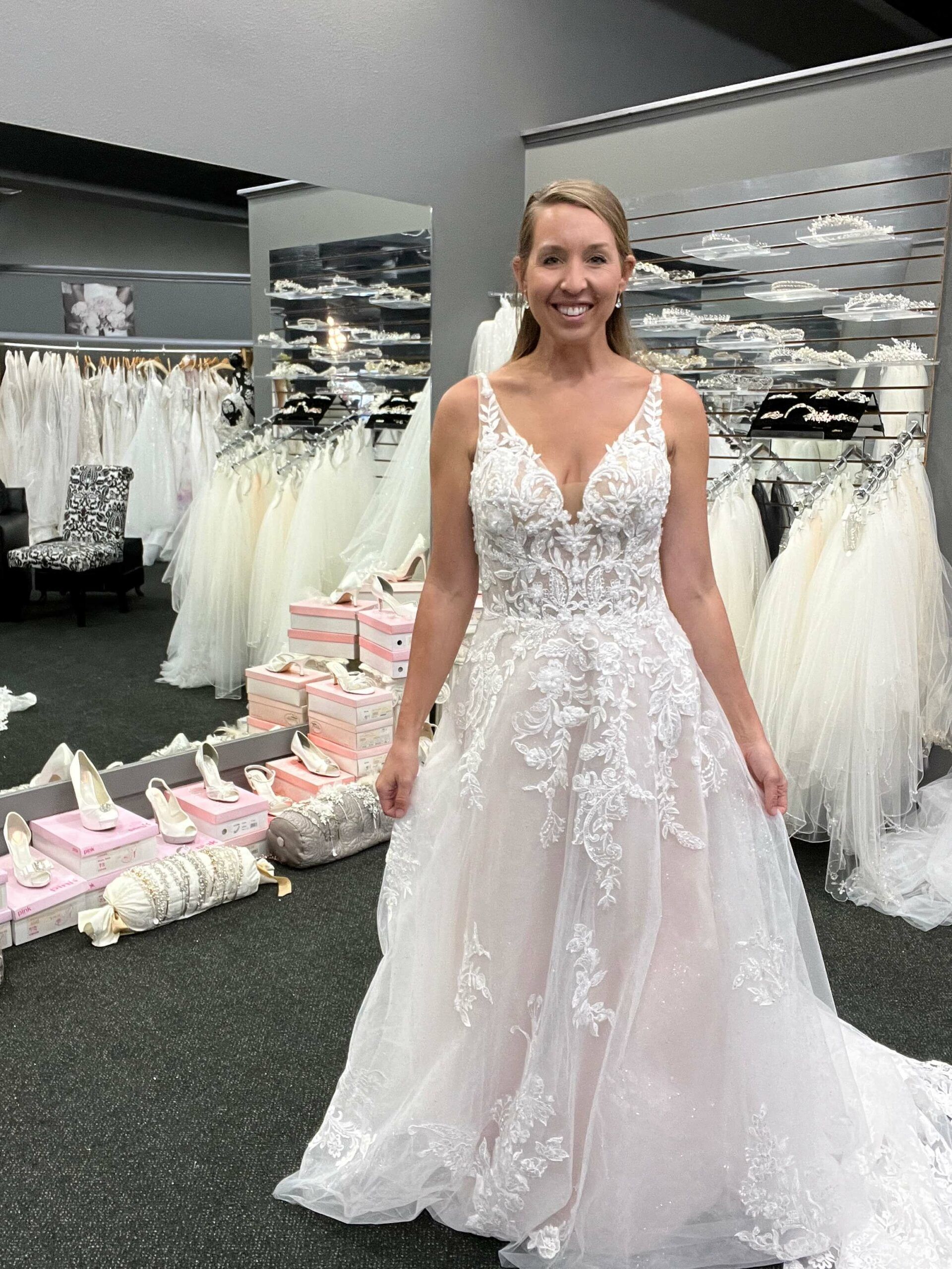 Woman smiles in a white wedding dress in a bridal shop, surrounded by gowns and shoes.