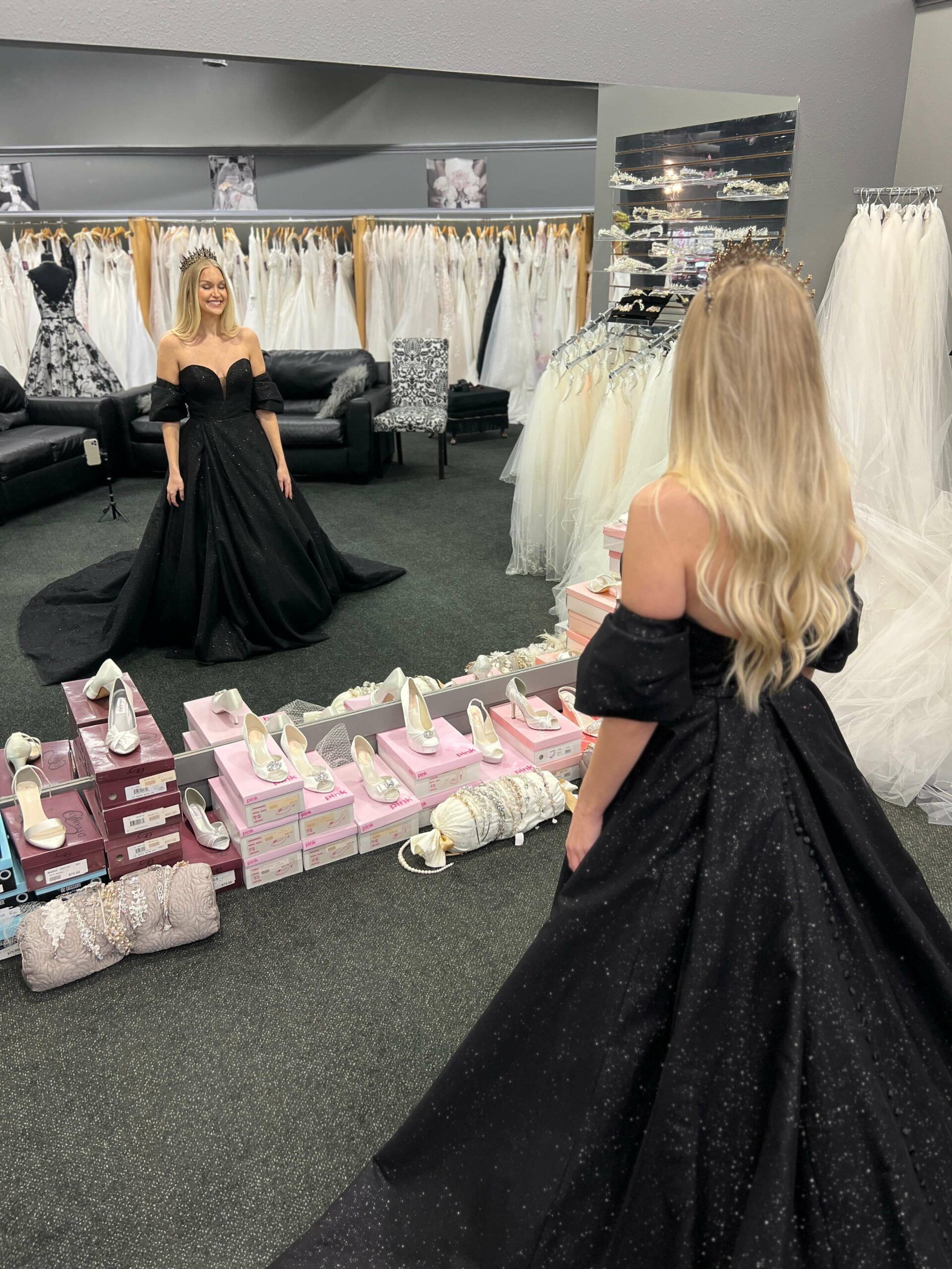 Woman in black gown admiring herself in a mirror, surrounded by wedding dresses and shoes in a bridal shop.