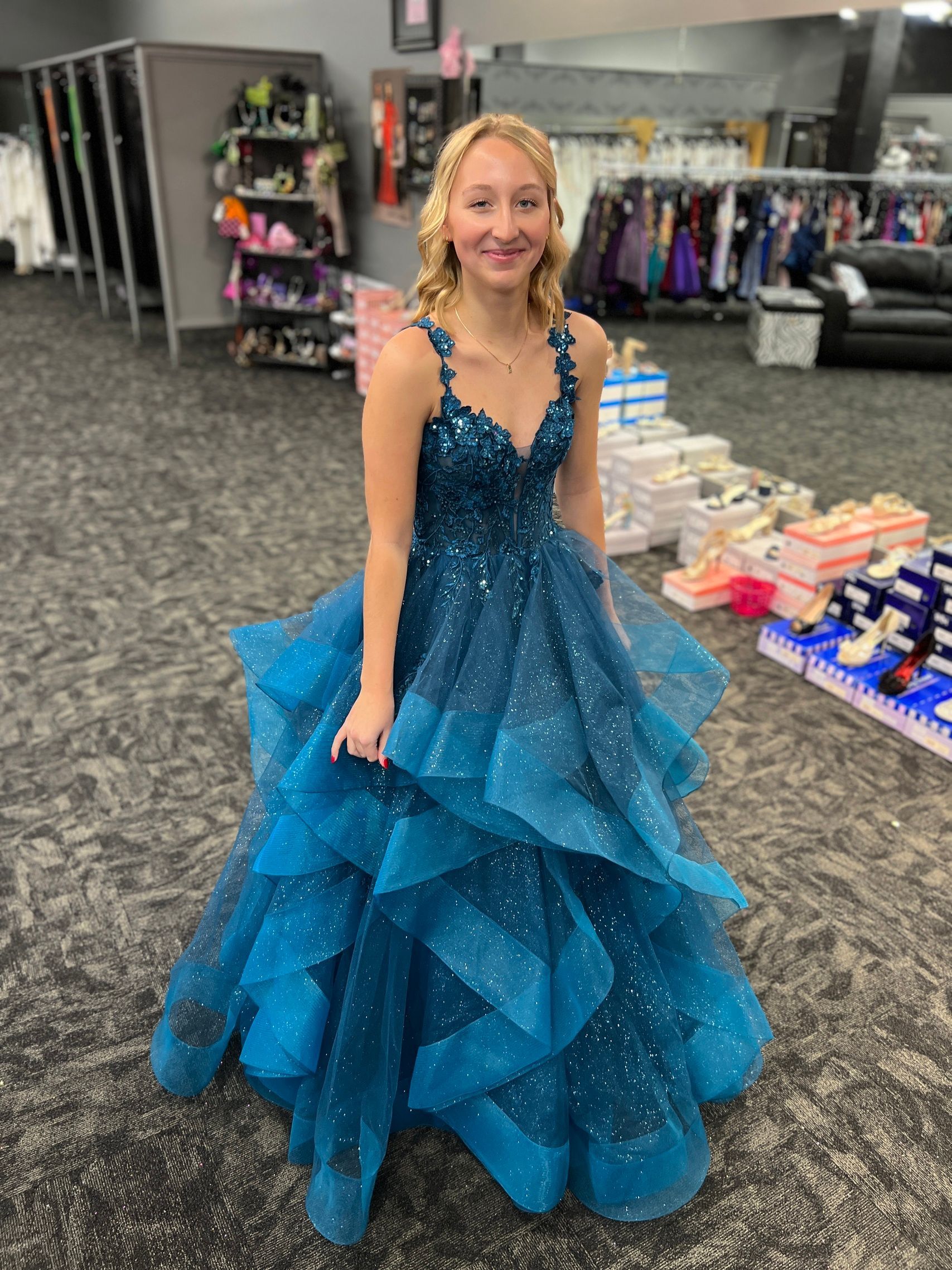 Blonde teen in teal sparkly prom dress, holding the skirt, smiles in a store.