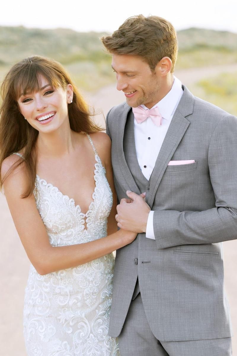Bride and groom smiling outdoors. The groom wears a gray suit with a pink bow tie. The bride is in a white lace dress.
