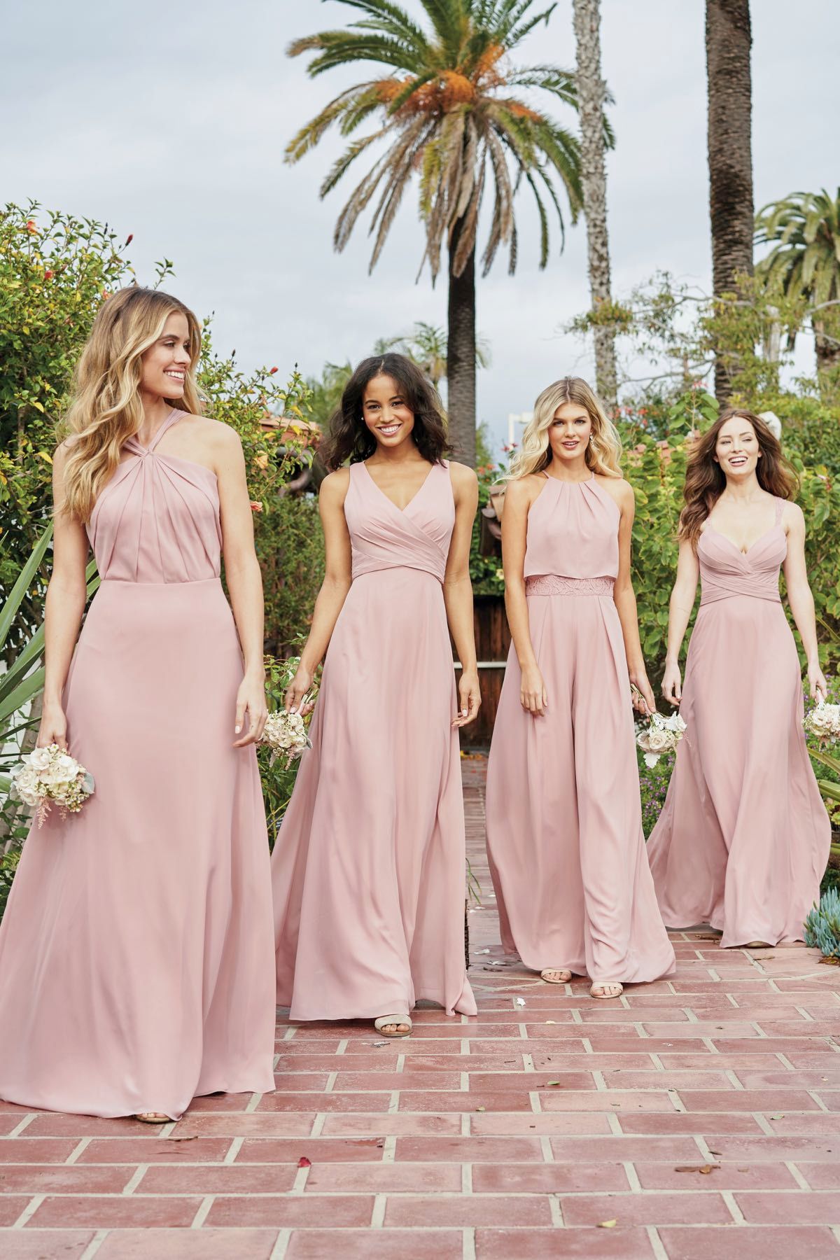 Four women in pink bridesmaid dresses walking on a brick path, smiling.