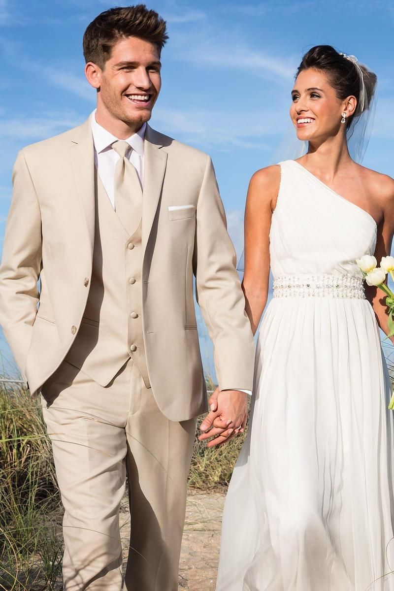 Couple in wedding attire hold hands, walking outdoors. Man in tan suit, woman in white one-shoulder gown, smiling.
