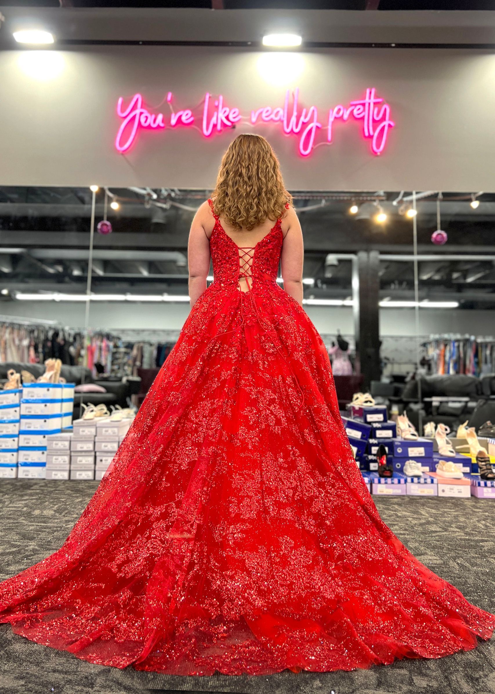 Woman in red sparkly gown, back to camera, in dress shop with neon sign.