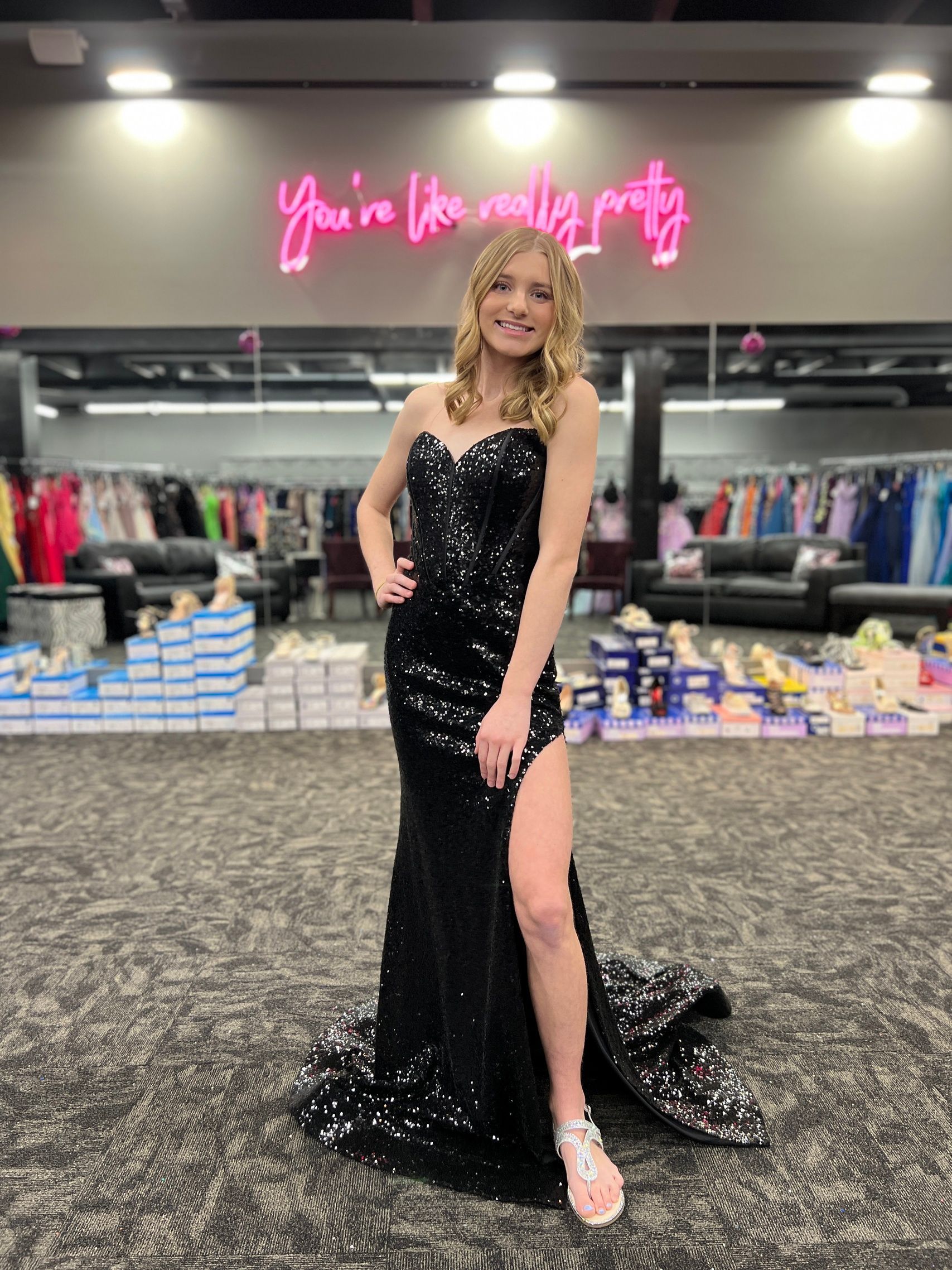 Woman in a black sequin gown poses in a formal wear store; a neon sign reads,