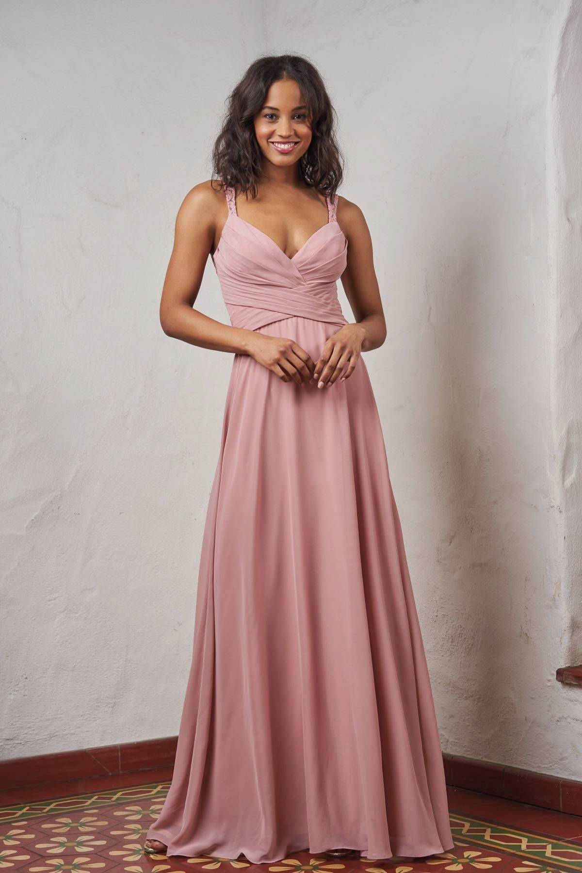 Woman in a dusty rose-colored gown, smiling, standing in a corner with textured walls.