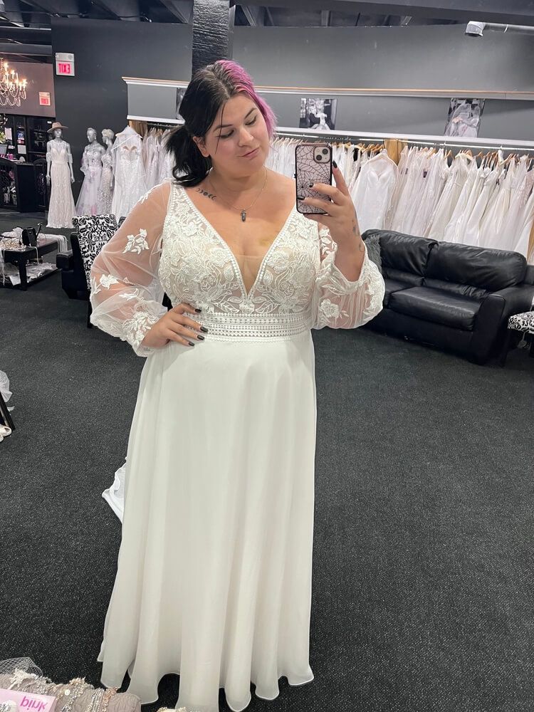 Woman in wedding dress, taking selfie in bridal shop. Dress has sheer sleeves, deep v-neck.