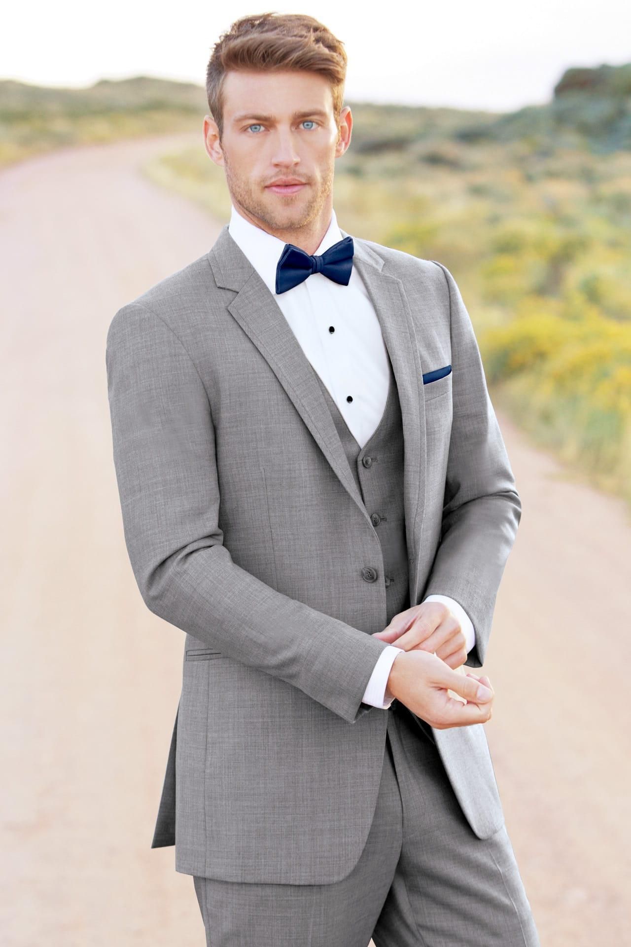Man in grey suit with navy bow tie, standing on a dirt road, looking at the viewer.
