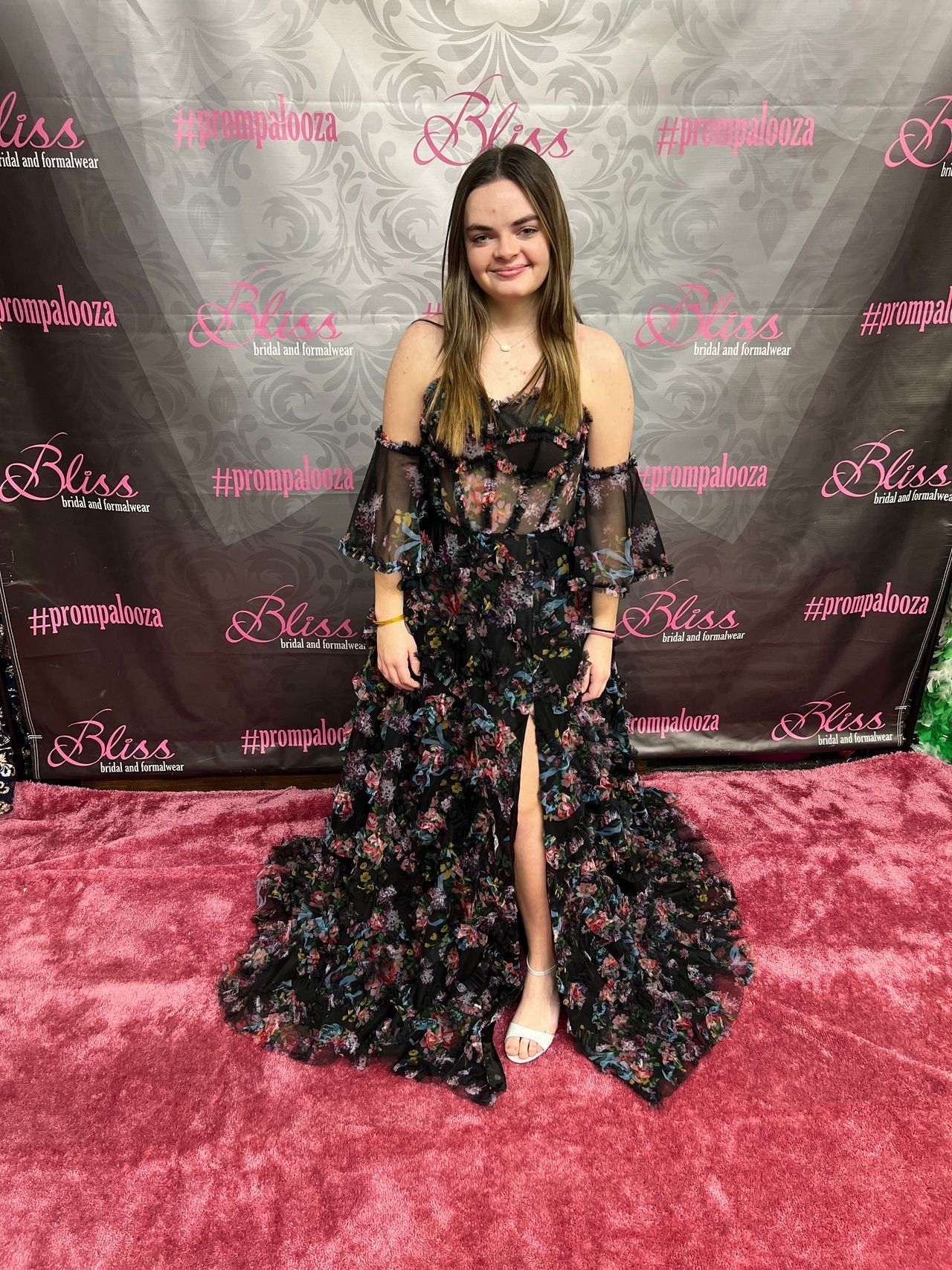 Woman in floral off-the-shoulder gown stands on a pink carpet in front of a backdrop with a logo.