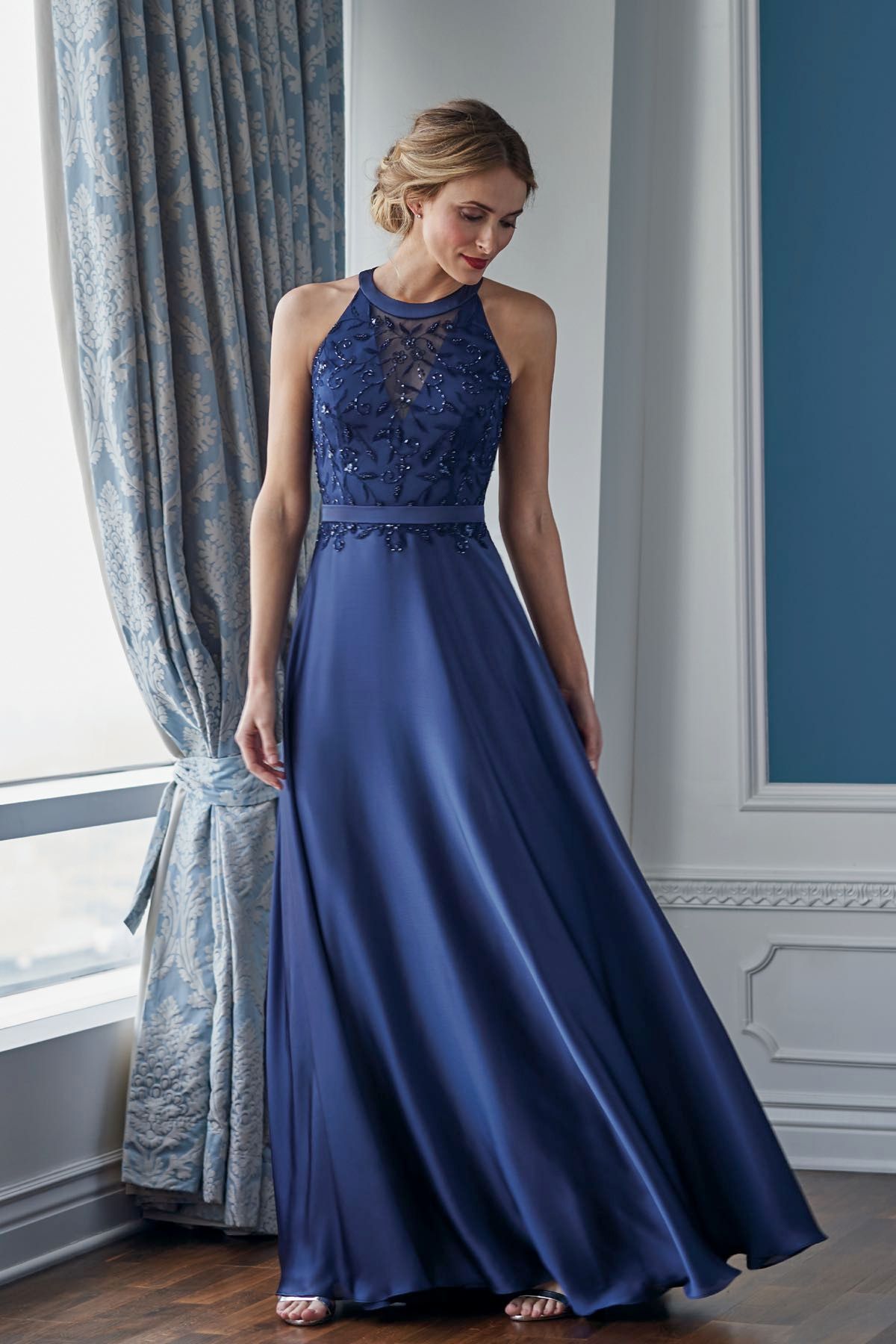 Woman in a navy blue formal gown with embellished bodice stands near a curtained window.