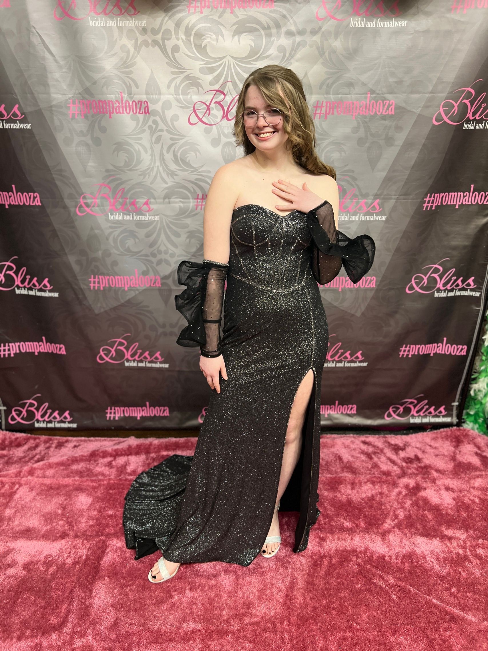 Woman in sequined black gown with a high slit, posing on a pink carpet in front of a banner.