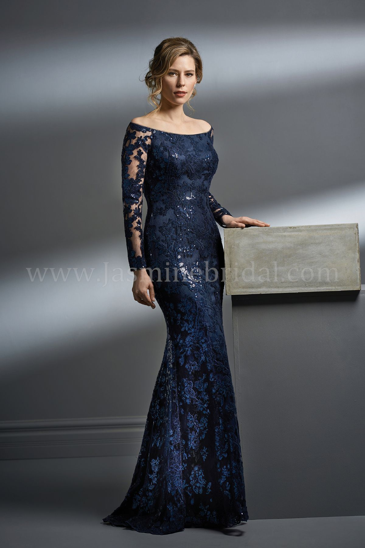 Woman in a navy blue, long-sleeved, off-the-shoulder gown with lace detailing, posing in front of a gray wall.