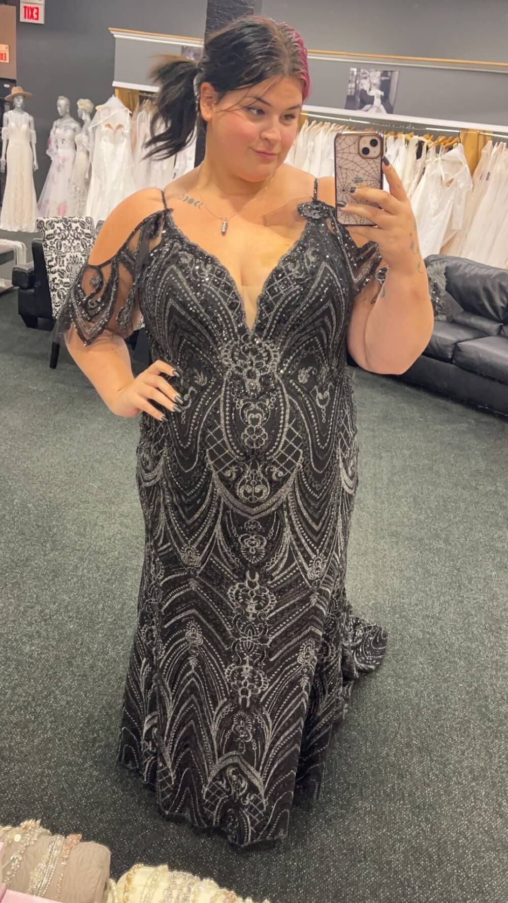 Woman in a black and silver formal gown in a bridal shop, taking a selfie.