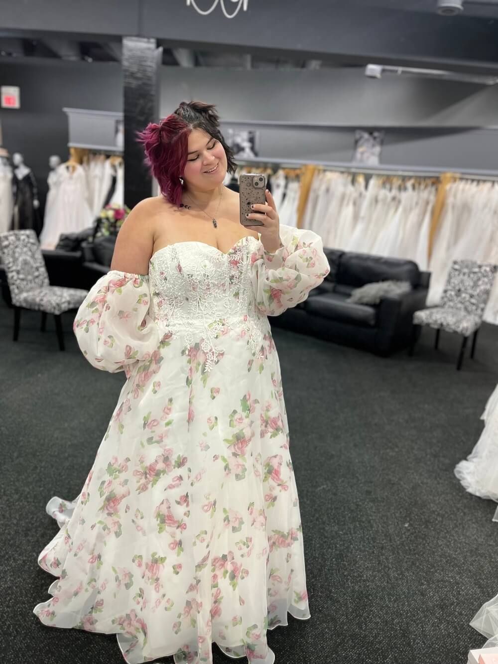 Woman in floral wedding dress takes a mirror selfie in a bridal shop.