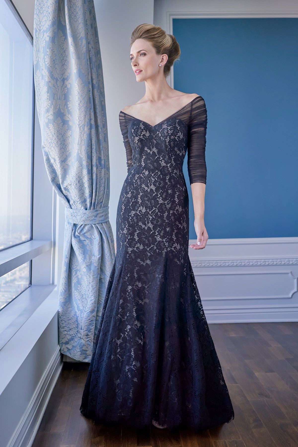 Woman in a navy lace gown, looking out a window. She's standing near a blue wall and curtain.