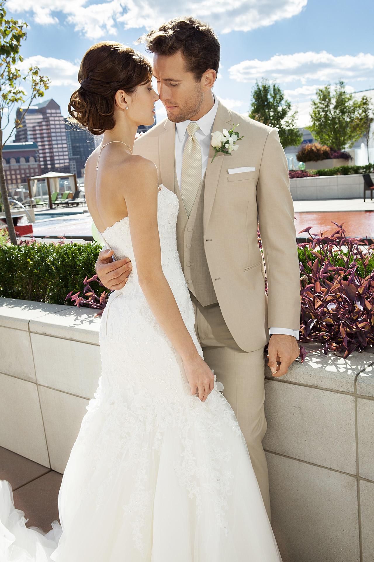 Couple in wedding attire embrace outdoors. Bride in strapless gown, groom in tan suit.