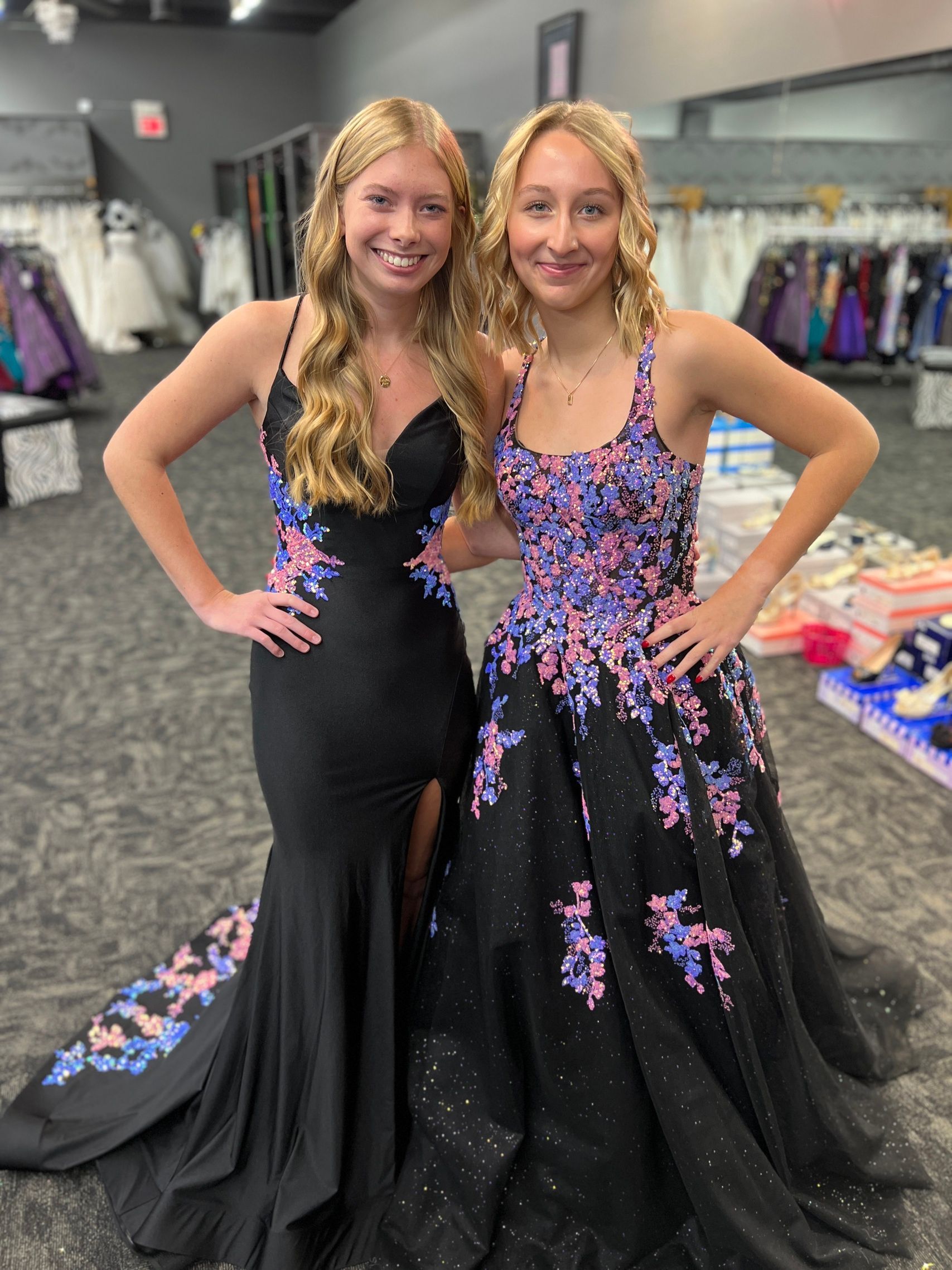 Two women in prom dresses, smiling, inside a dress shop.