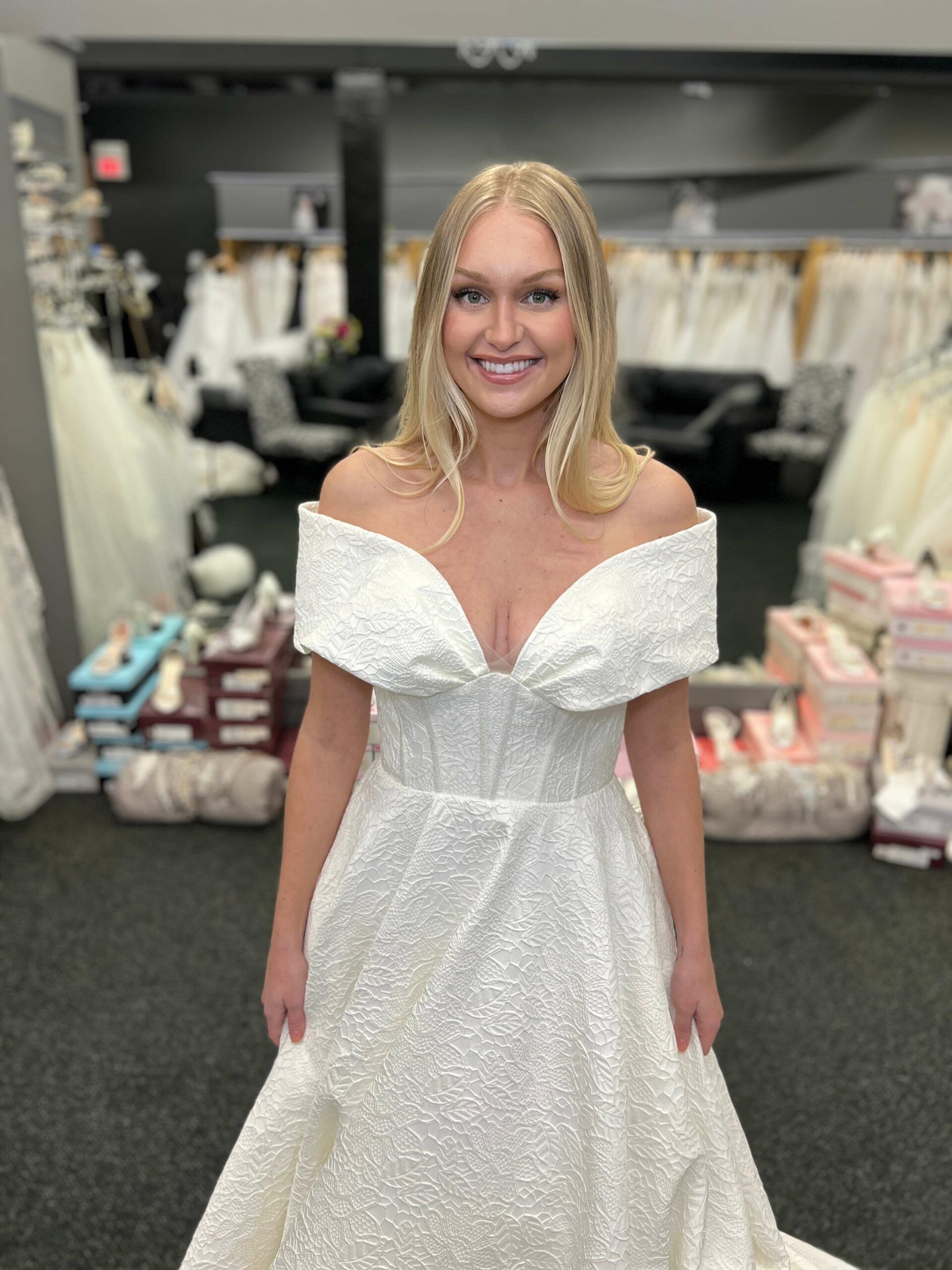 Woman in a white off-the-shoulder wedding dress smiles in a bridal shop.