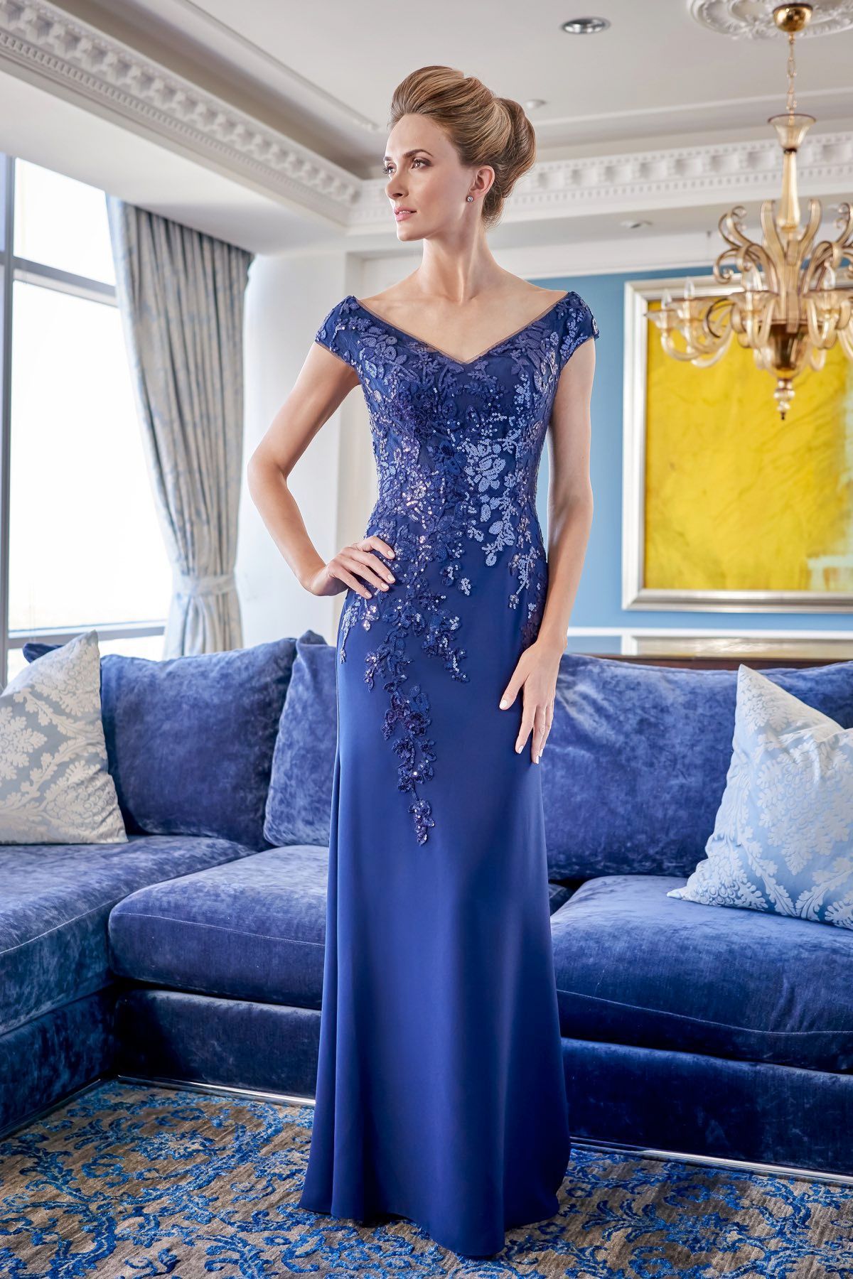 Woman in a blue evening gown with embellished bodice stands indoors, hand on hip.