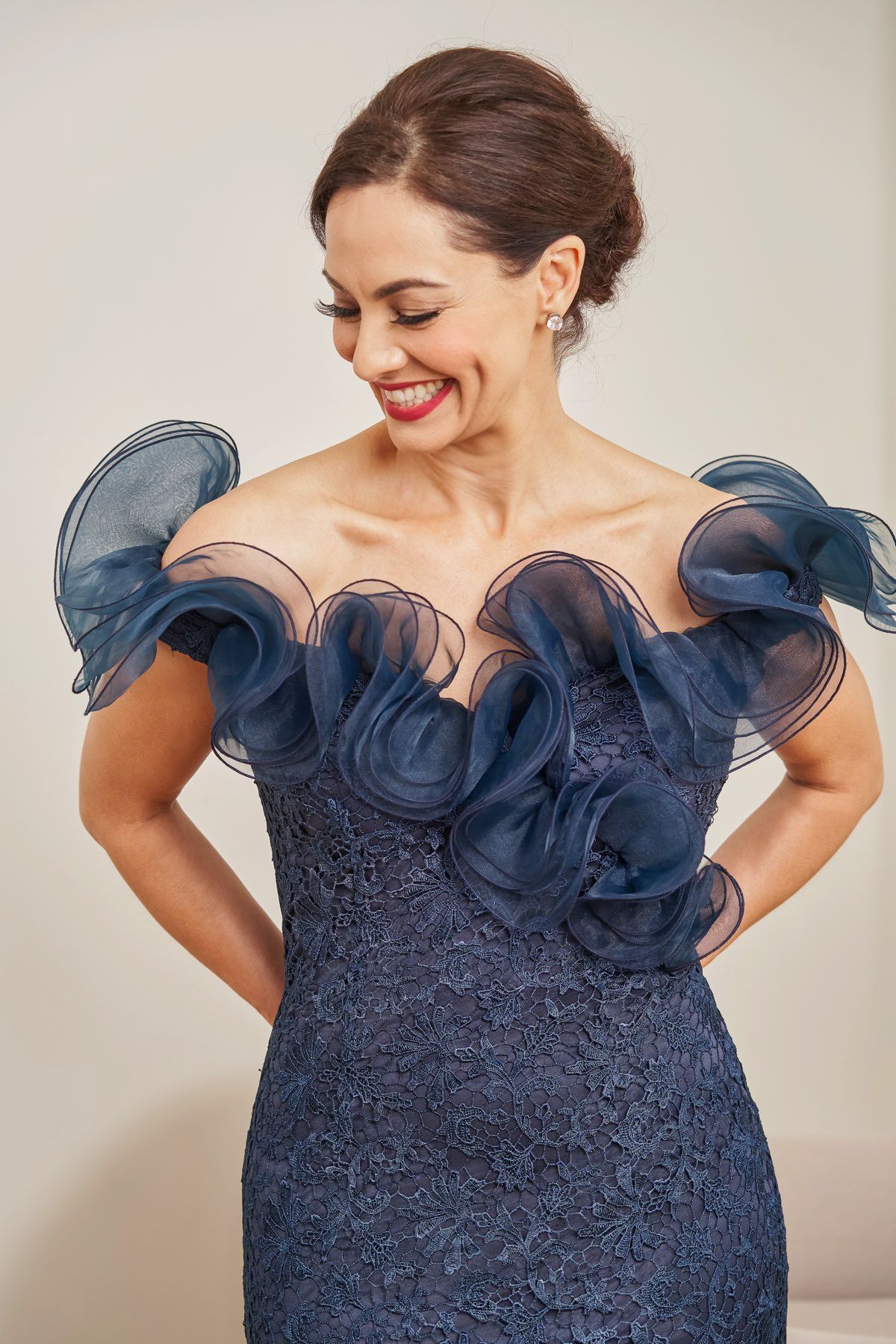 Woman in a navy off-the-shoulder gown, smiling. Hair in updo, hands at side. Light background.