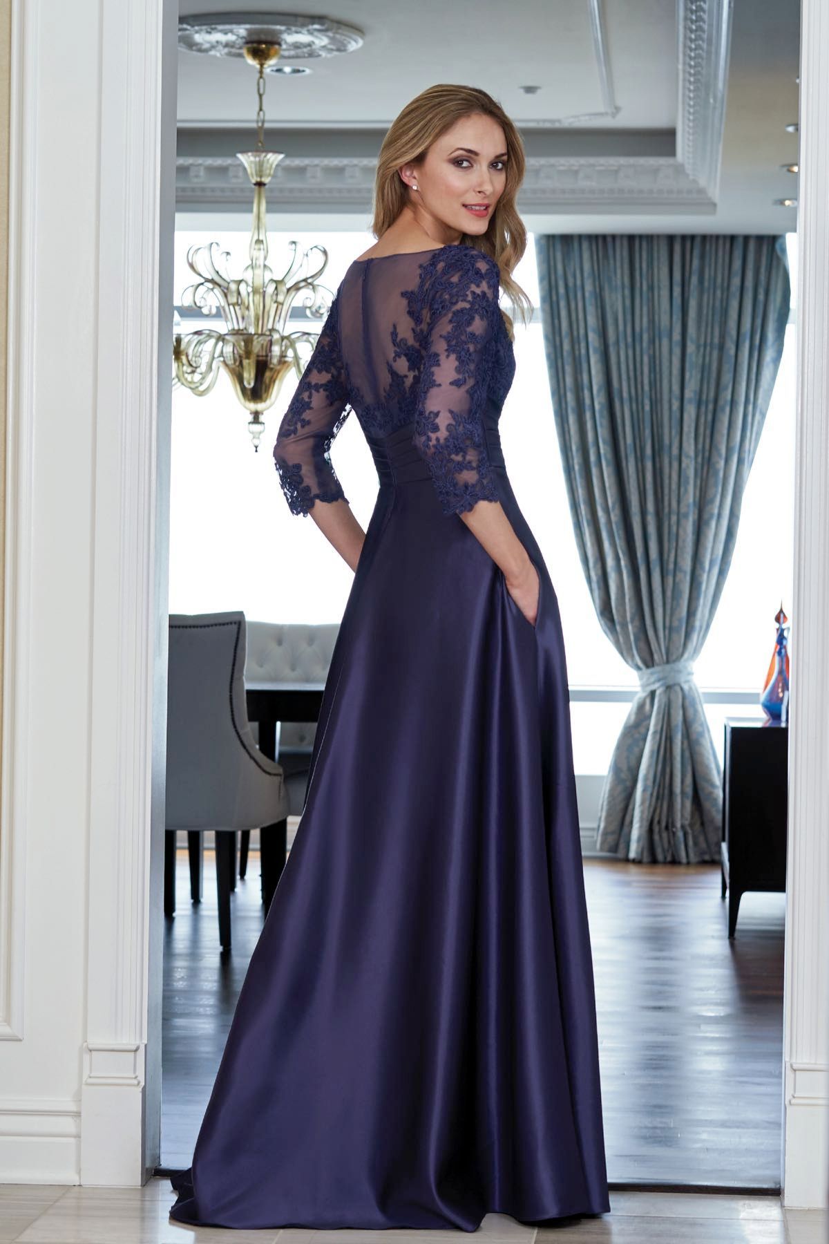 Woman in a navy blue satin gown with lace sleeves and back, standing in a doorway.