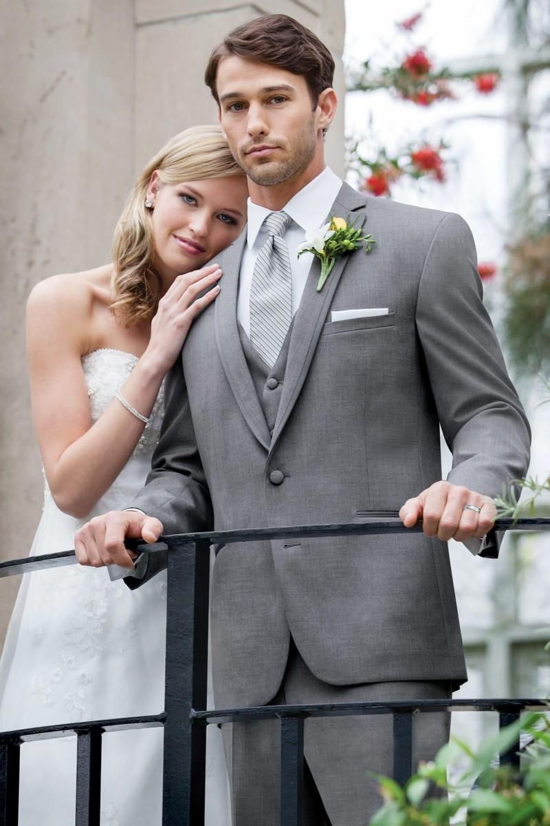 Bride and groom in formal wear on a balcony, man in gray suit, woman in white dress, both looking at camera.