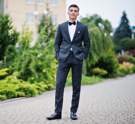 Man in a dark suit with a bow tie, standing on a brick path, hands in pockets.