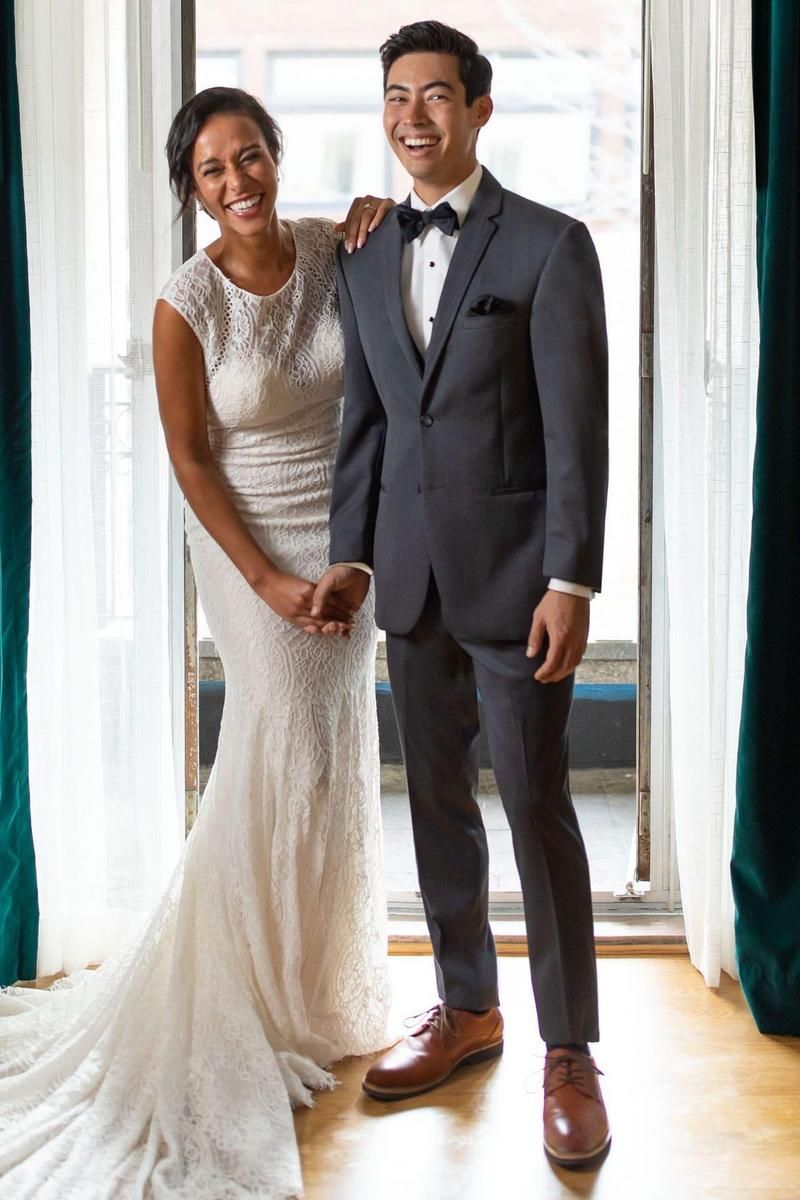 Bride and groom smiling, holding hands, in formal attire. Standing near window with teal curtains.