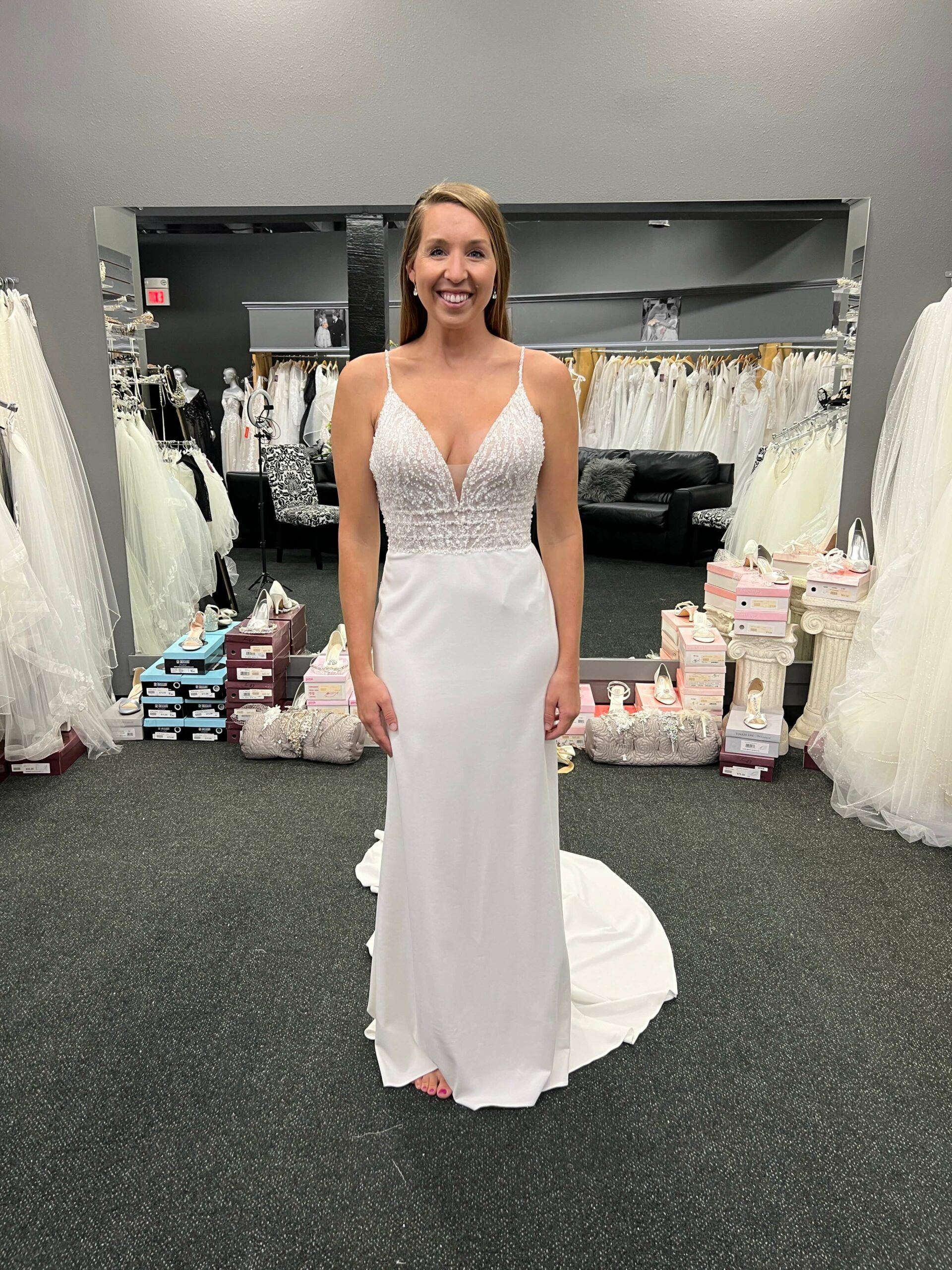 Woman in white wedding dress, smiling in front of a mirror in a bridal shop.