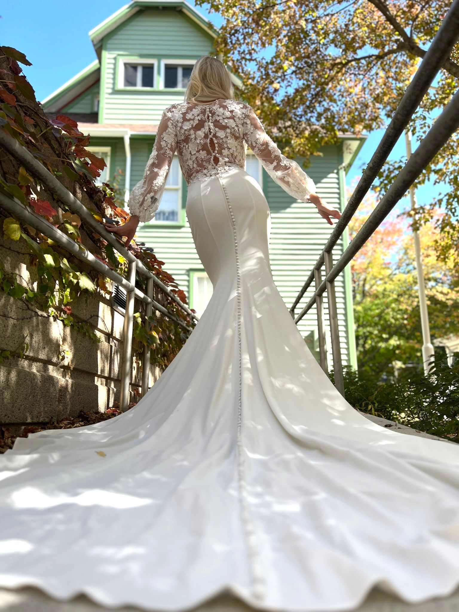 Woman in white wedding dress with long train, standing outside a house.