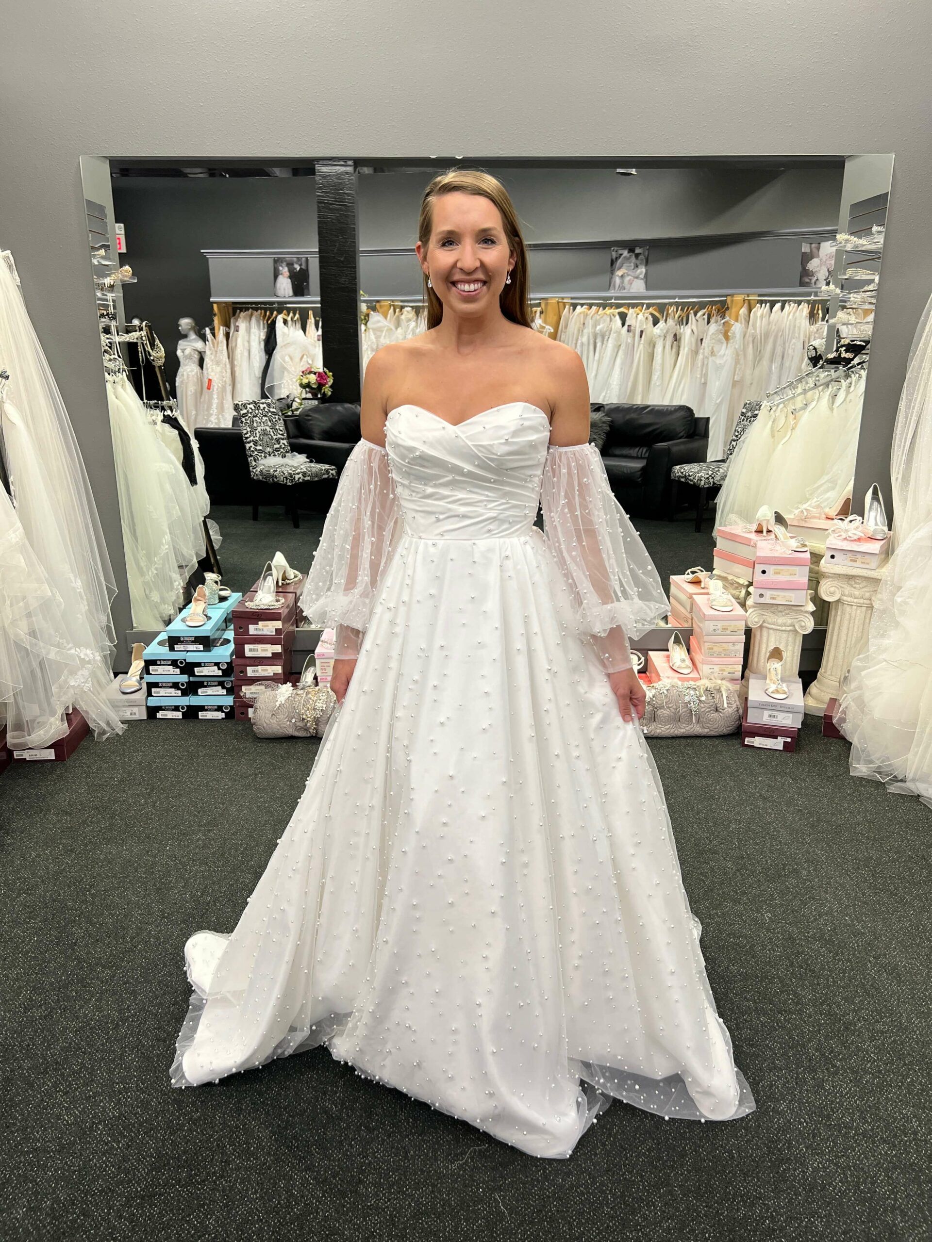 Woman in a white off-the-shoulder wedding dress with long, sheer sleeves smiles in a bridal shop.