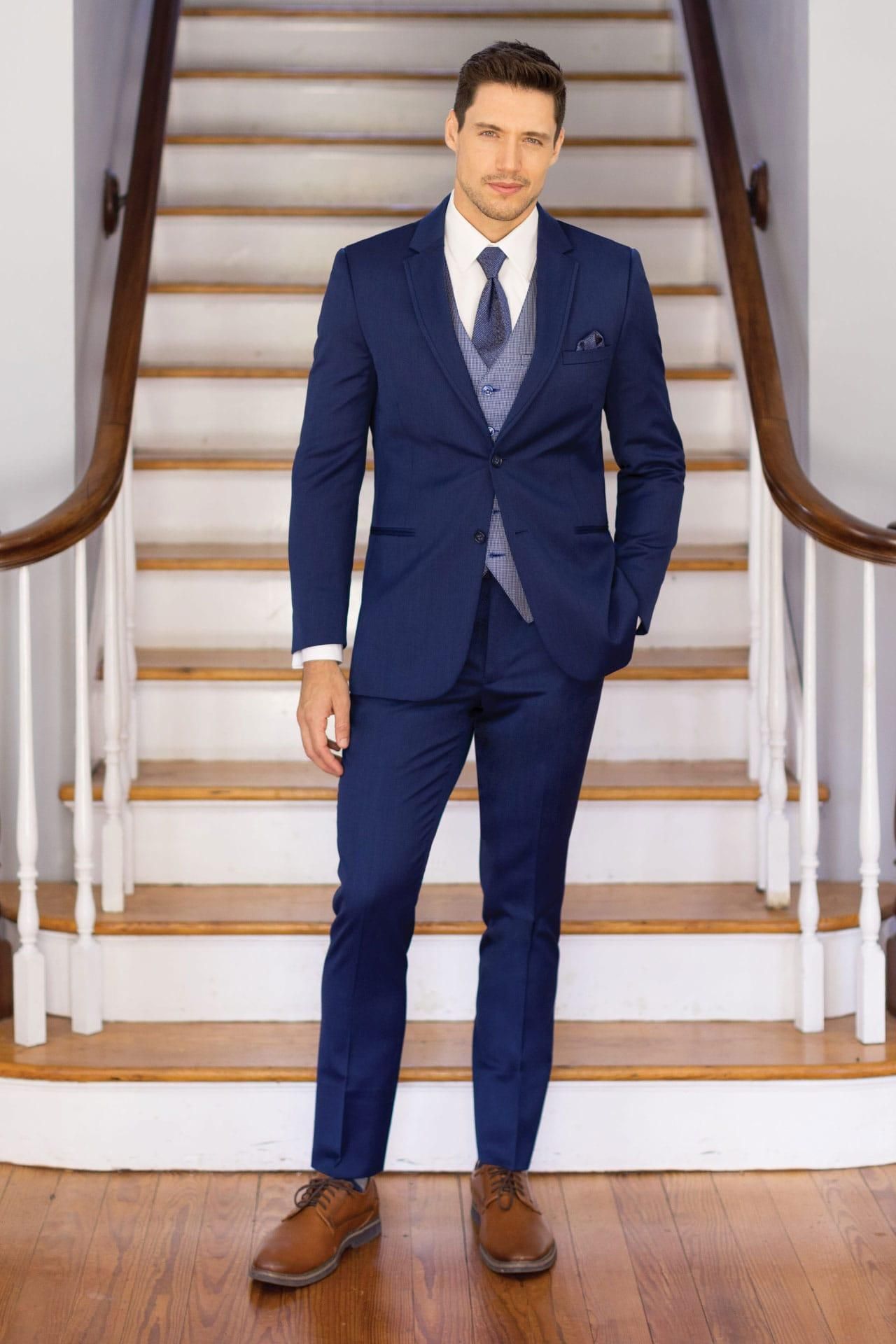 Man in a navy blue suit and vest, brown shoes, stands on a wooden floor in front of a staircase.