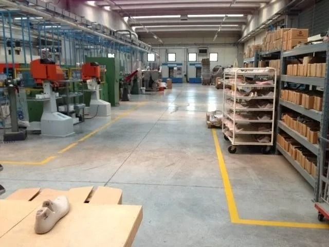 Interno di una fabbrica con macchinari, forme per scarpe e scaffali. Le linee gialle delimitano i percorsi pedonali.