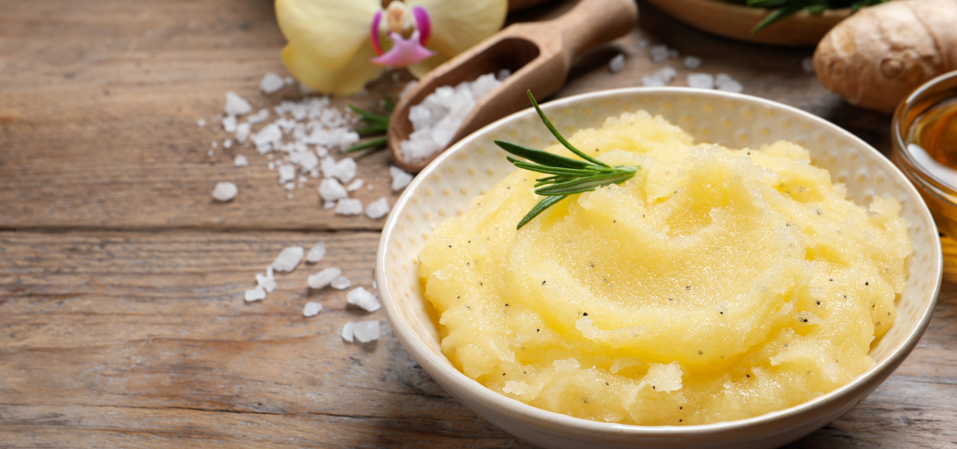 Bowl of creamy mashed potatoes topped with rosemary on a rustic wooden table, with ingredients nearby