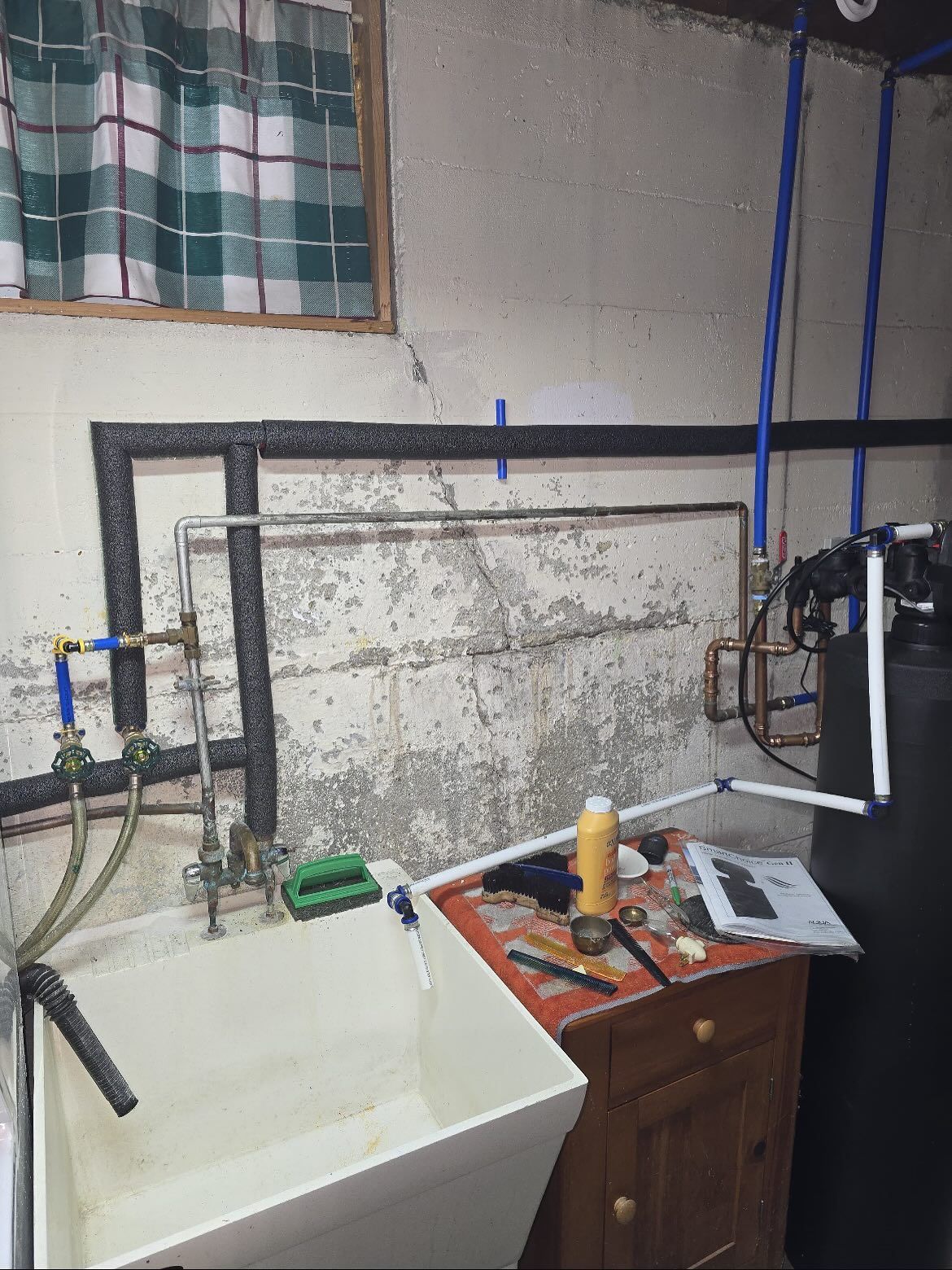 Small utility room with sink, pipework, tools on a table, and a small white appliance against a worn wall.