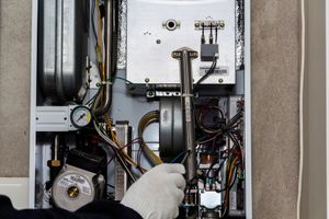 Technician in gloves inspecting furnace wiring with a wrench inside an open HVAC unit.