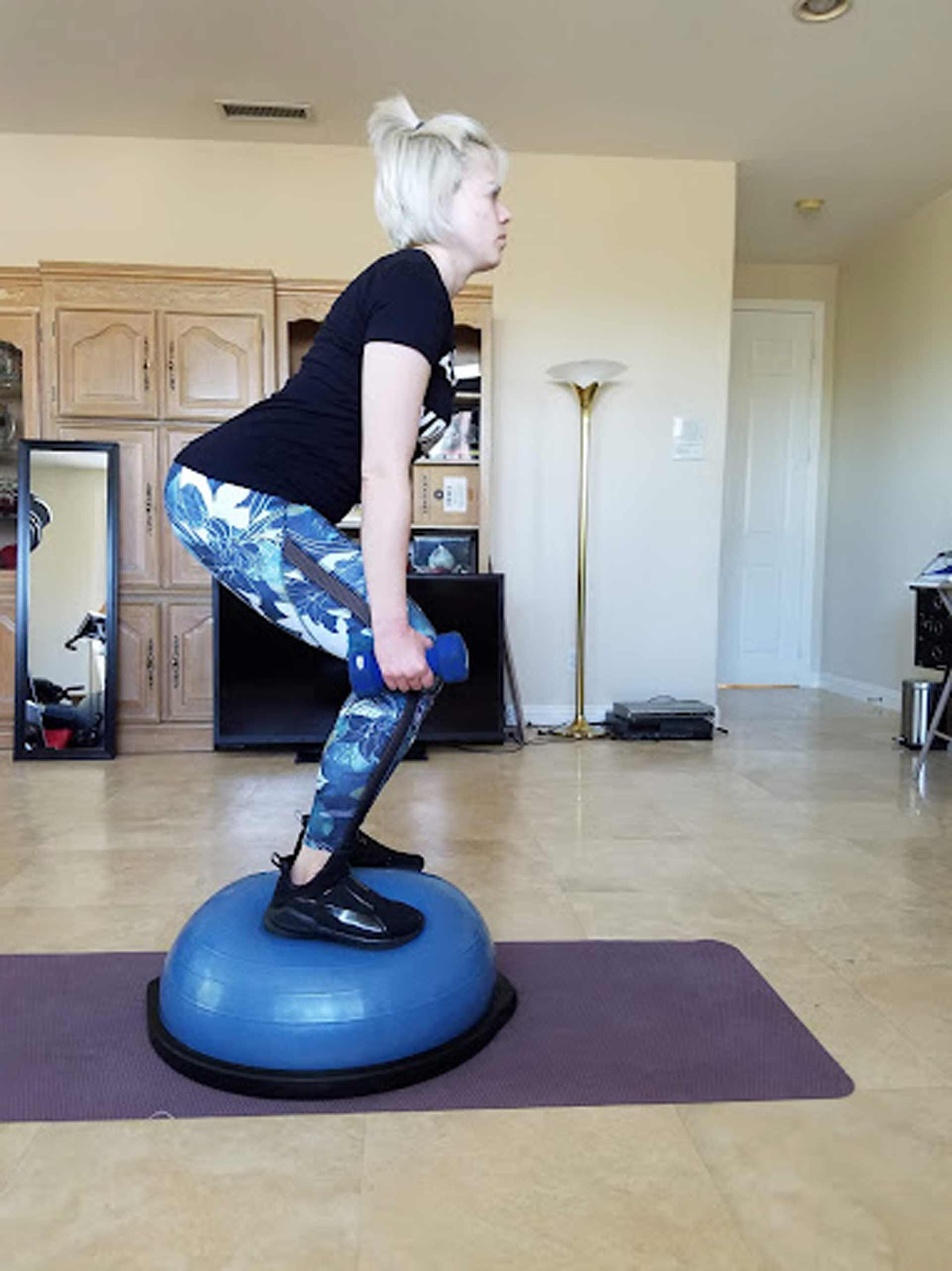 A woman is squatting on a blue exercise ball while holding a dumbbell.